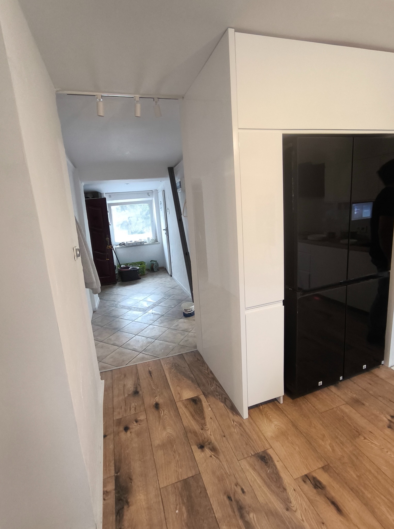 Nowoczesna kuchnia z białymi szafkami i czarną lodówką side-by-side. Widoczny fragment korytarza z płytkami i odnowioną podłogą. Świeże malowanie ścian.