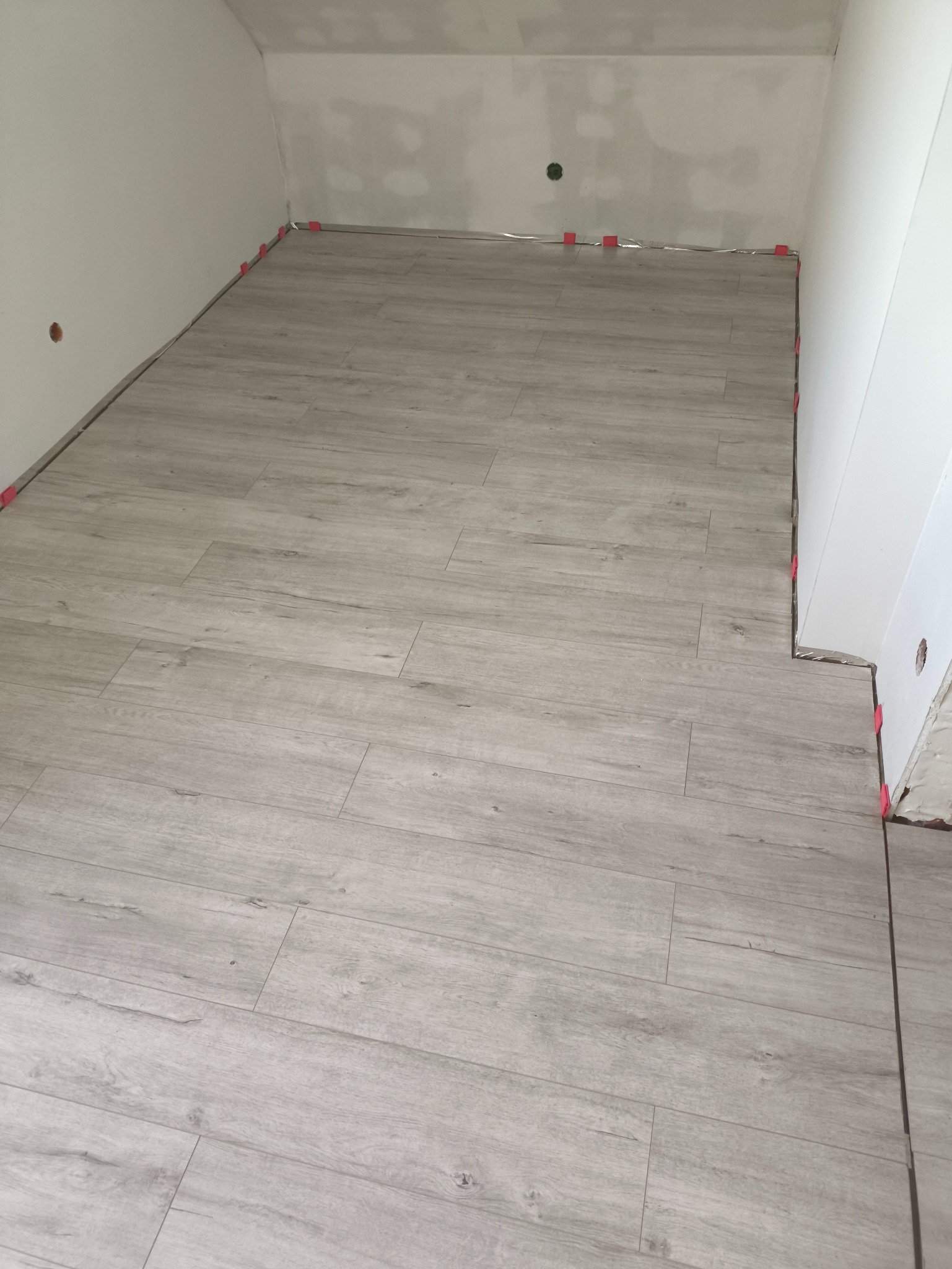 Ułożone panele podłogowe w odcieniach szarości, z widocznymi elementami dystansowymi przy ścianach. Ściany przygotowane do dalszych prac wykończeniowych.
