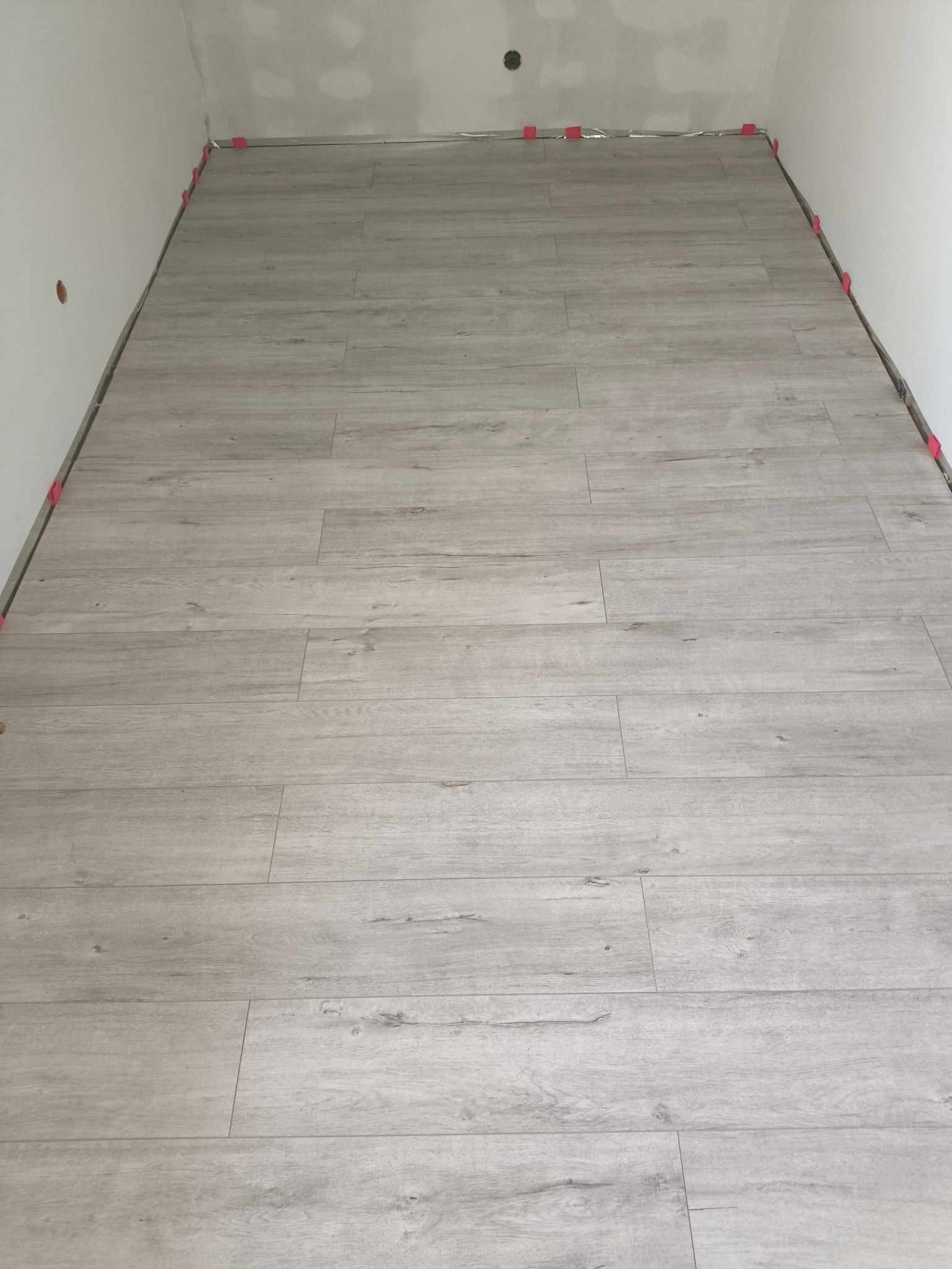 Jasne panele podłogowe ułożone w pomieszczeniu, widoczne czerwone kliny dystansowe przy ścianach. Imitacja drewna z delikatnym wzorem słojów.