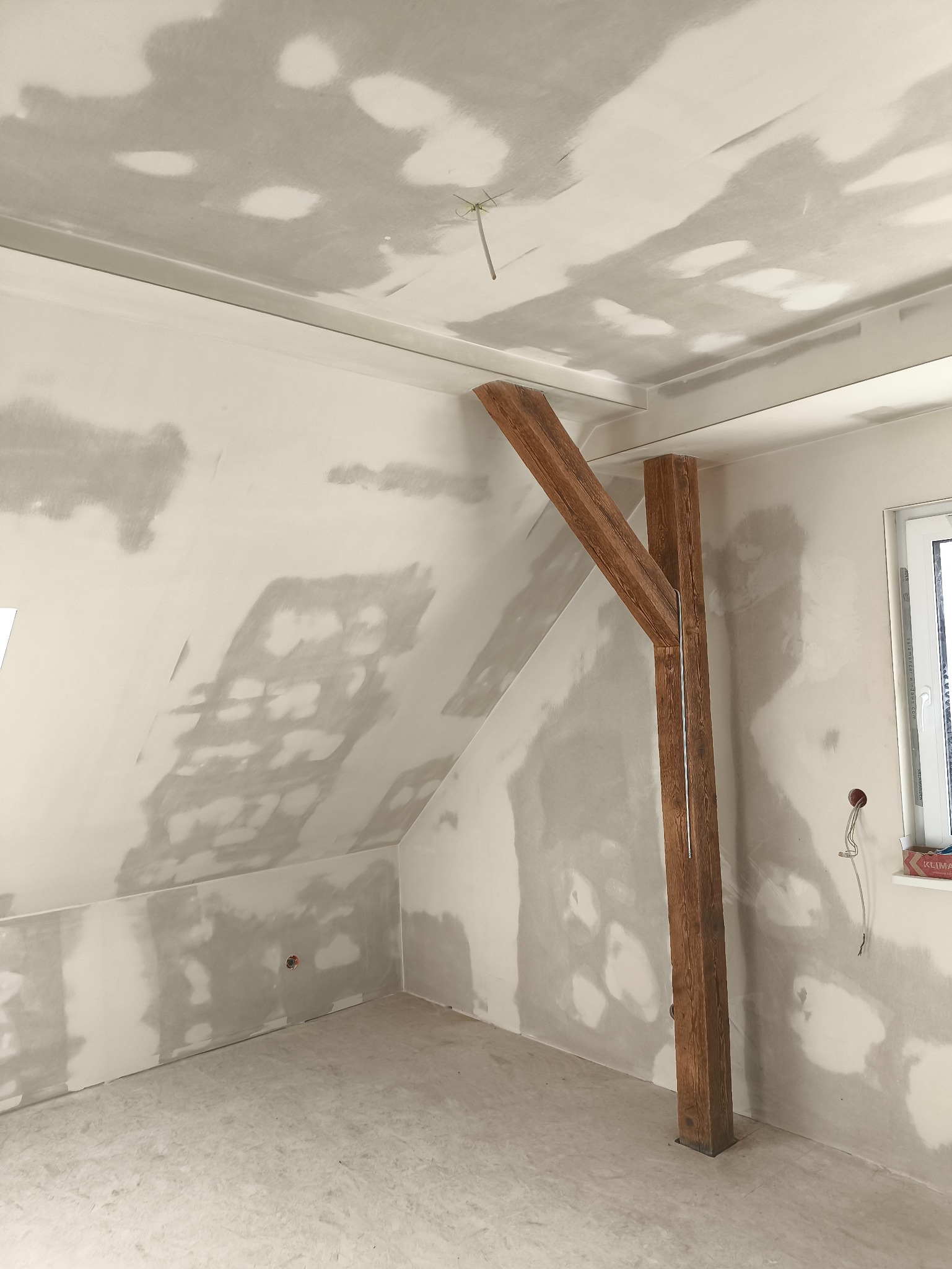 Poddasze w trakcie prac wykończeniowych: szpachlowane ściany, drewniana belka konstrukcyjna, okno. Widoczne ślady po szpachlowaniu na ścianach i suficie.