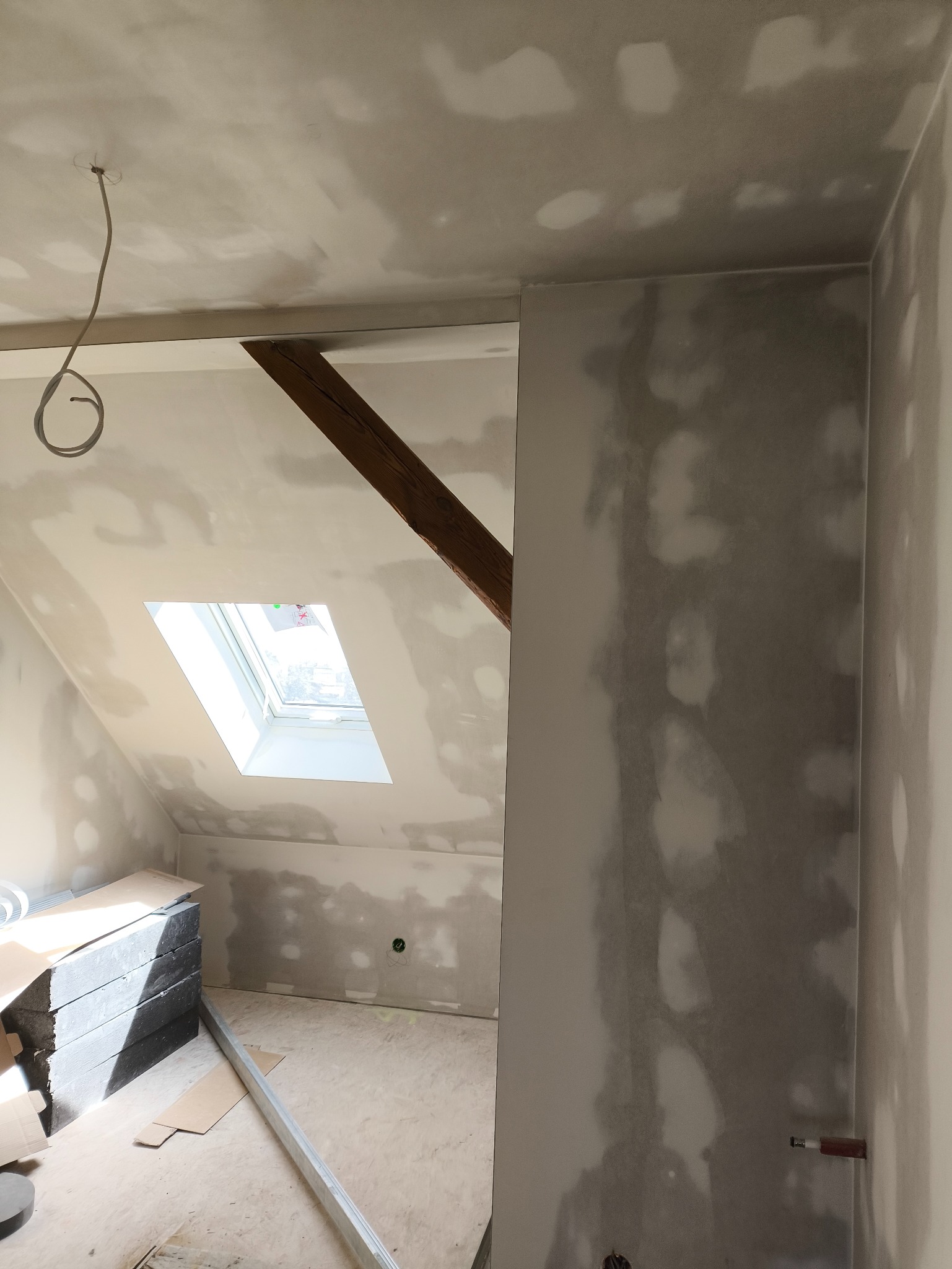 Poddasze w trakcie prac wykończeniowych: ściany przygotowane do malowania, widoczne łączenia płyt gipsowych, okno dachowe i drewniana belka stropowa.
