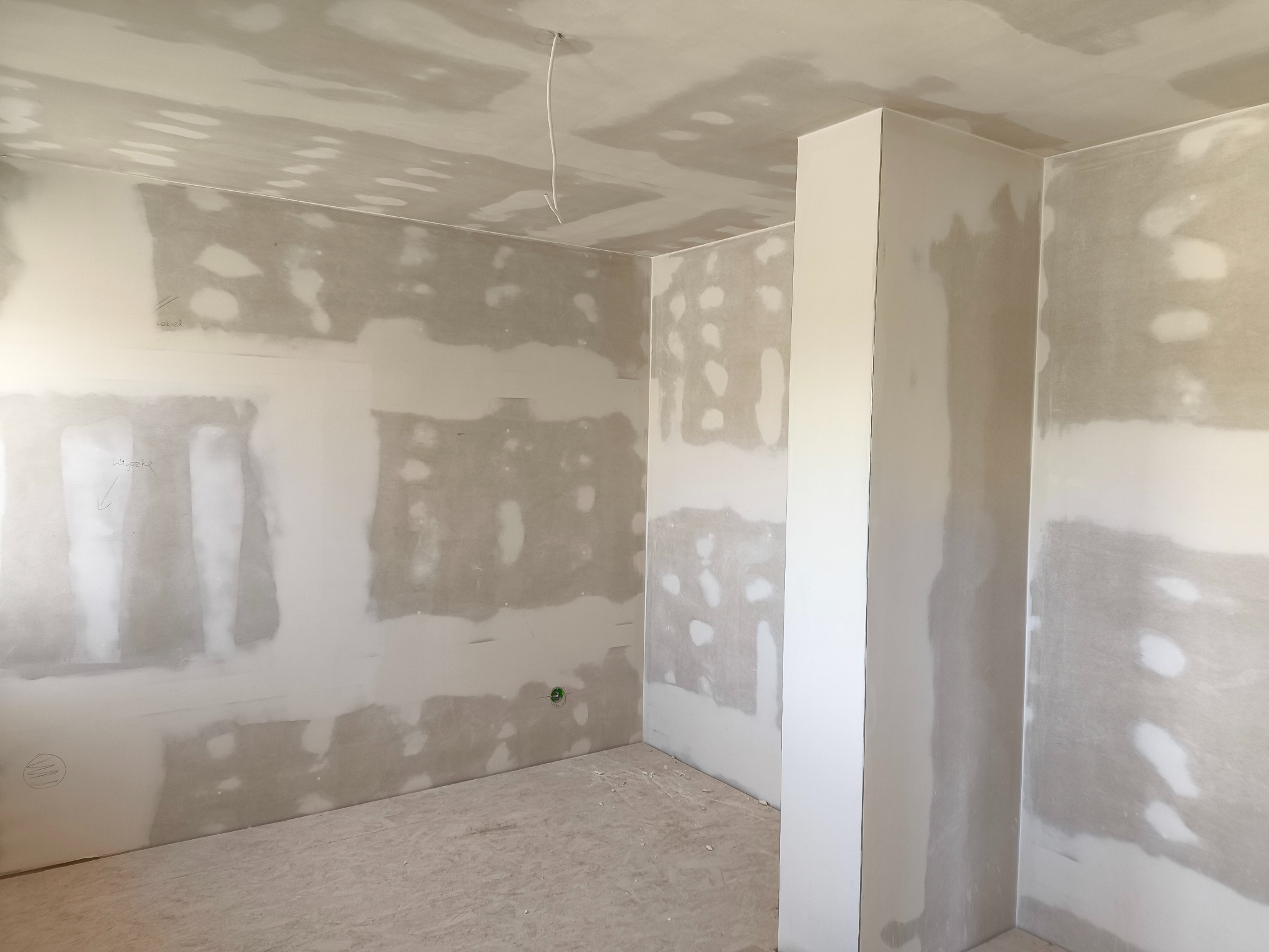 Wnętrze pokoju w trakcie prac wykończeniowych: ściany z płyt gipsowo-kartonowych, widoczne łączenia i szpachlowanie. Na suficie zwisający kabel elektryczny.