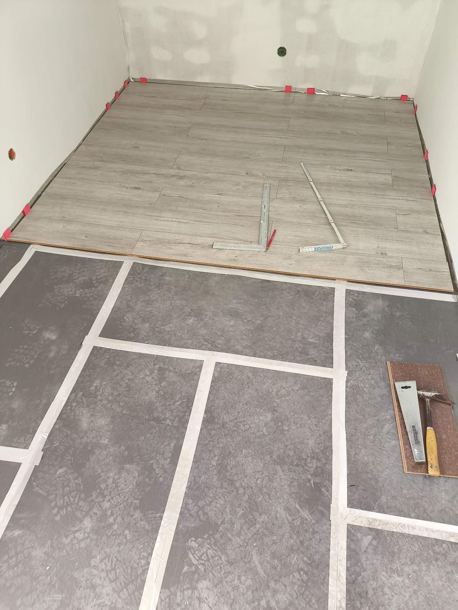Fragment układania paneli podłogowych w pomieszczeniu. Widoczne narzędzia, podkład i częściowo ułożone panele z dystansami przy ścianie. Ujęcie z góry.