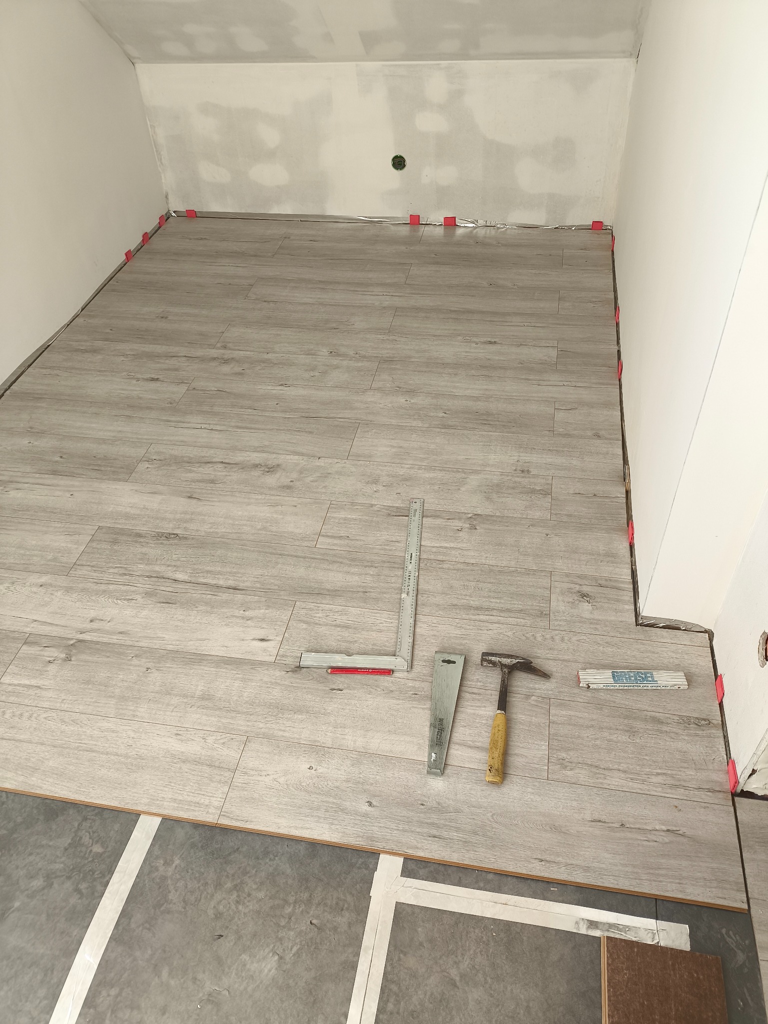 Układanie paneli podłogowych w pokoju na podkładzie, widoczne narzędzia: młotek, kątownik, listwa. Czerwone kliny dystansowe przy ścianach. Częściowo ułożona podłoga.