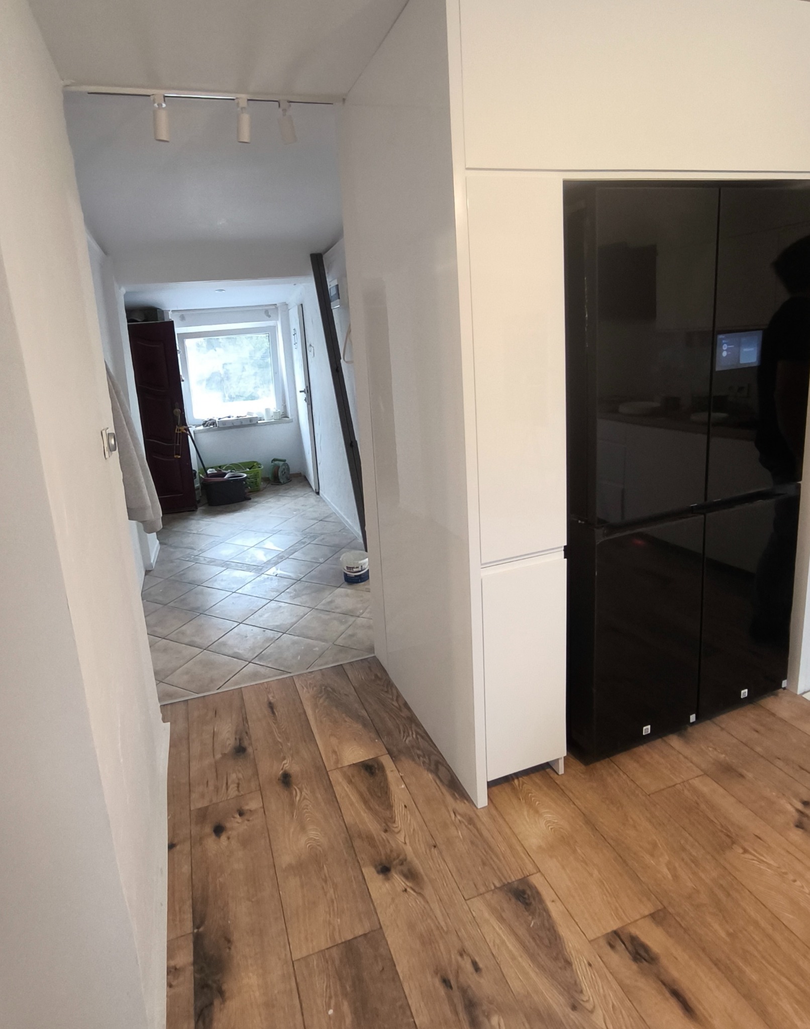 Jasne panele podłogowe w korytarzu z widokiem na okno. Ściany białe, oświetlenie sufitowe. Widoczne wiadro z farbą i narzędzia malarskie.