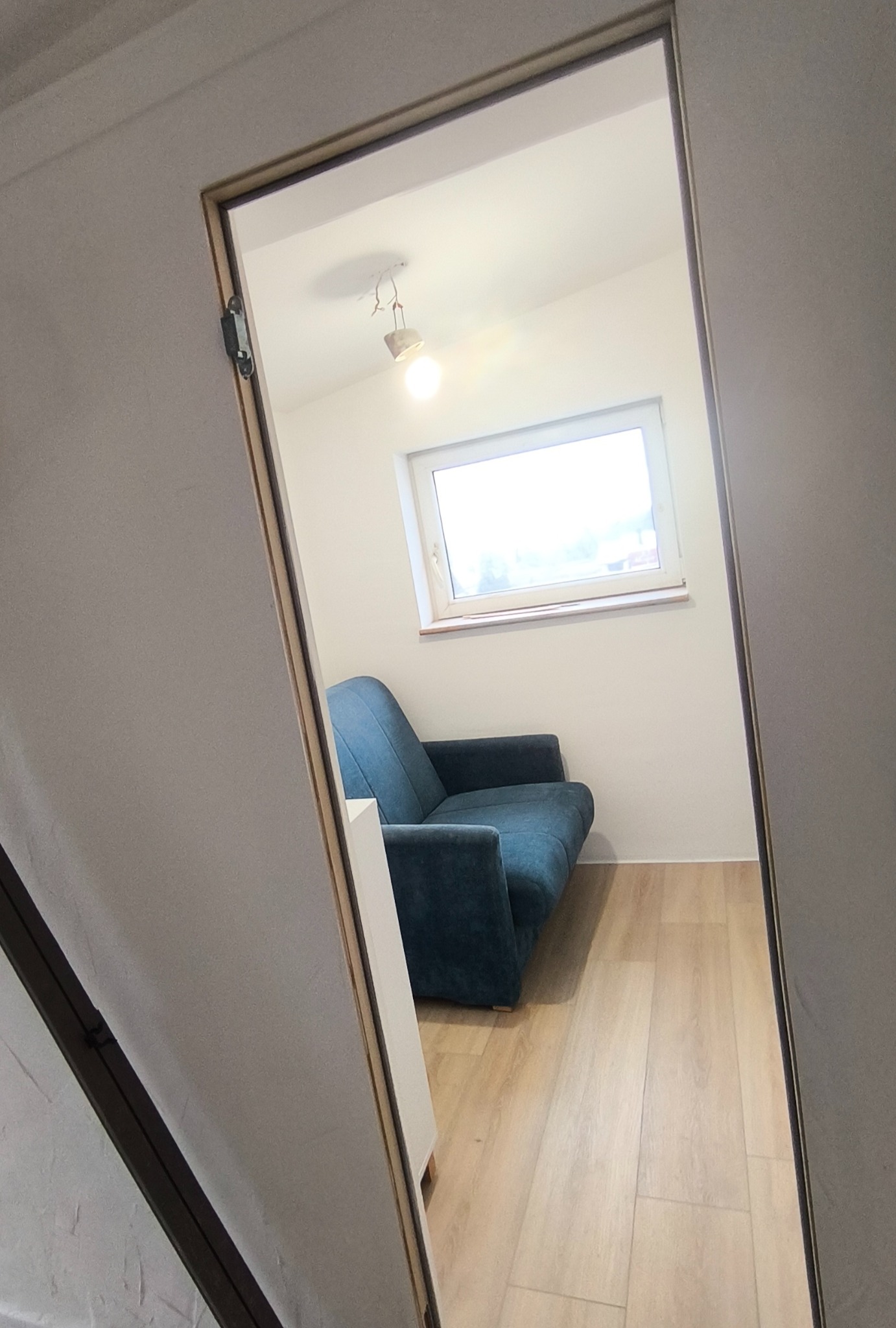 Ukończone wnętrze: błękitna sofa w pokoju z jasną podłogą, widoczne przez futrynę drzwi. Minimalistyczny design, świeże, jasne kolory, lampa sufitowa.