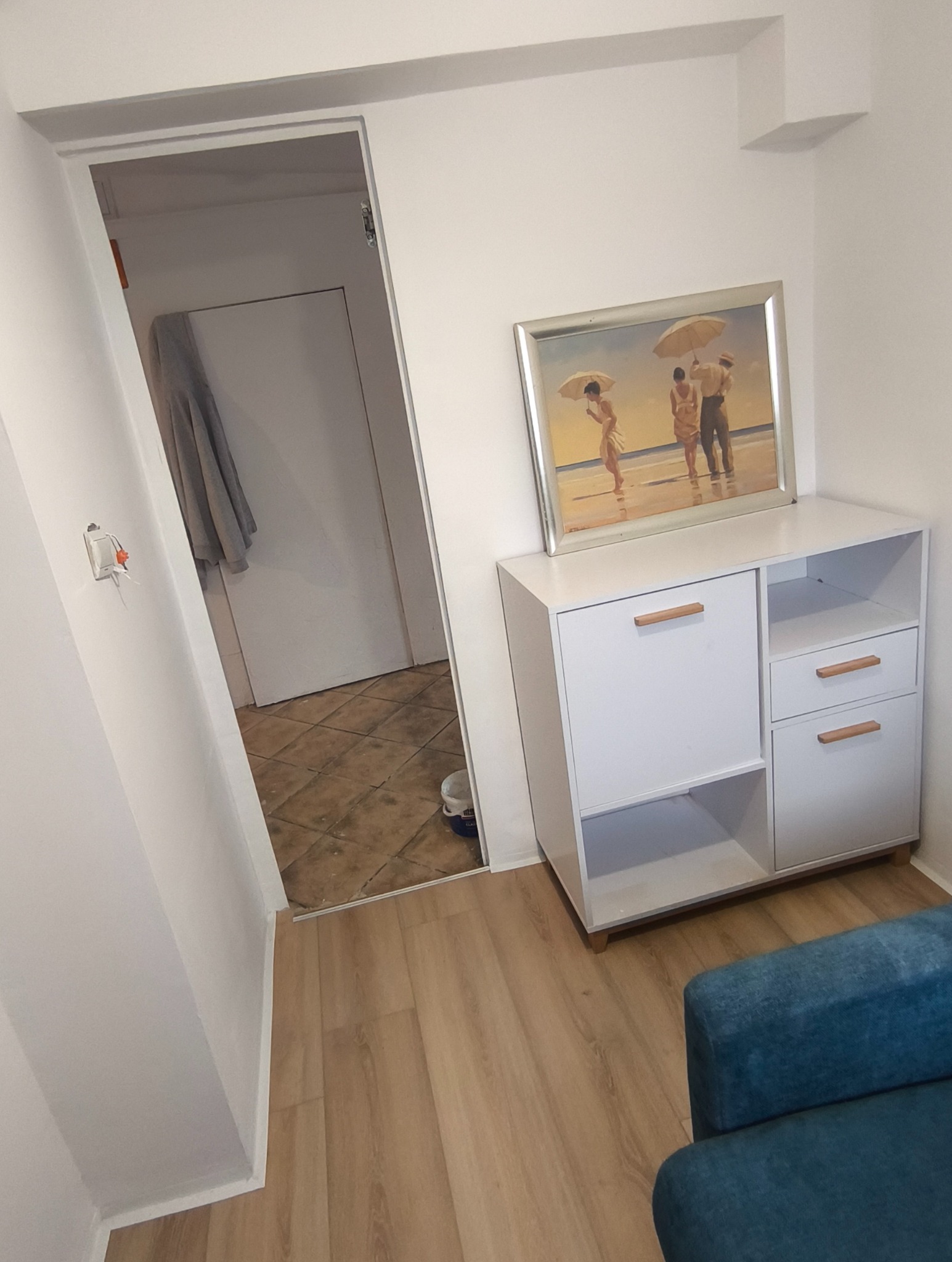 Biała komoda z obrazem plaży w pokoju z panelami podłogowymi i otwartymi drzwiami. Widoczny fragment niebieskiego fotela.