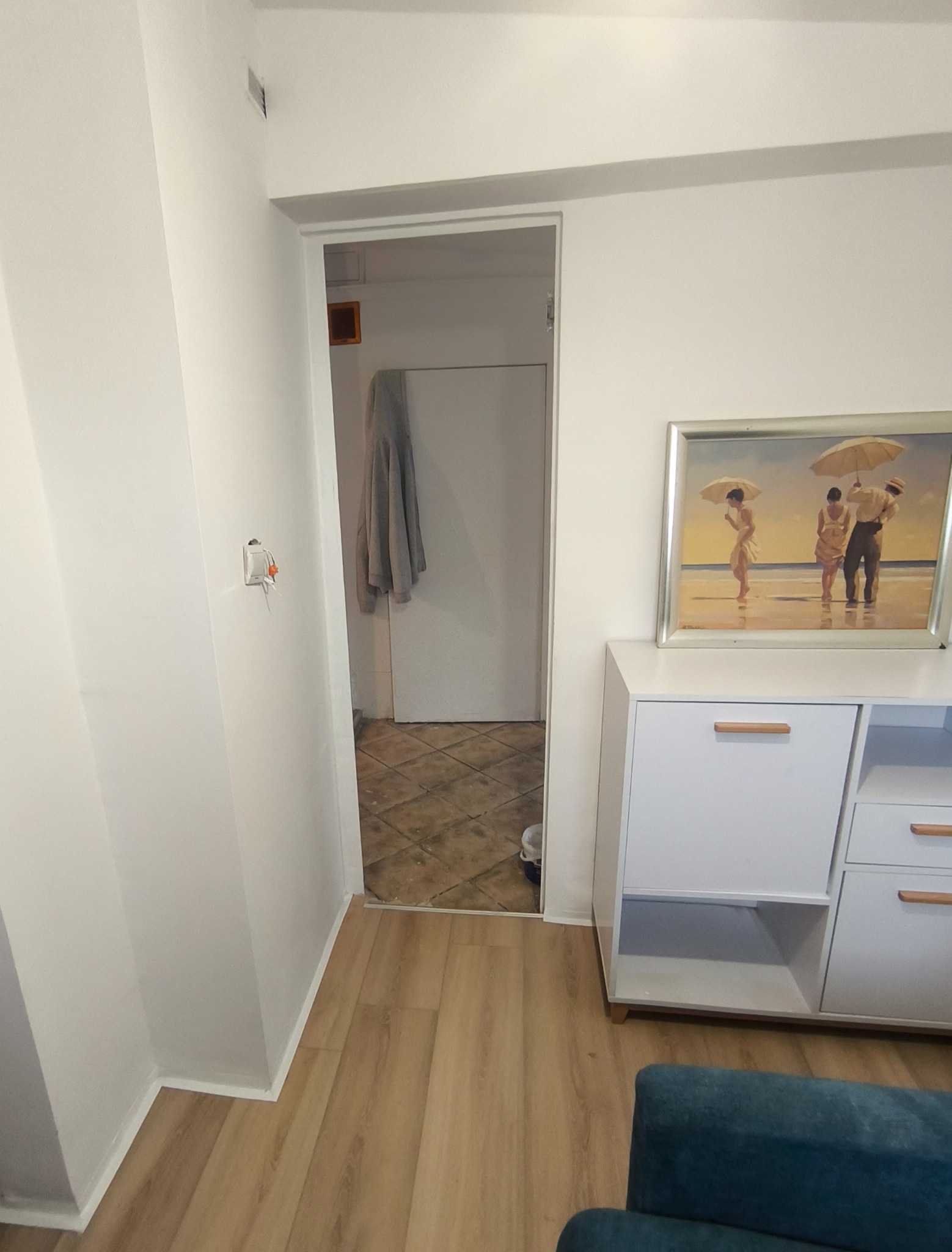 Wnętrze mieszkania po remoncie: białe ściany, jasne panele podłogowe, widoczny fragment szafki z obrazem. W tle otwarte drzwi do innego pomieszczenia z brązowymi płytkami.