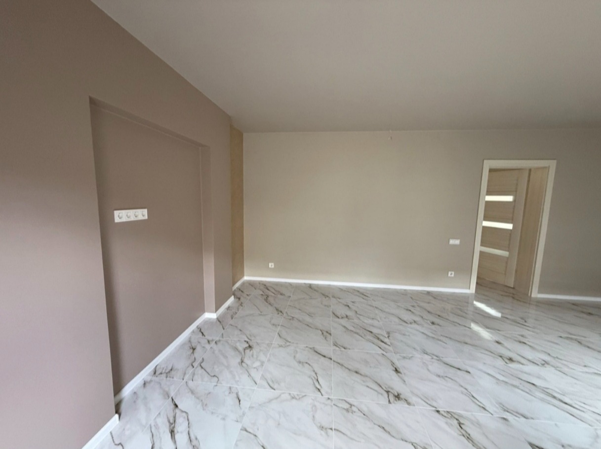 Jasne, świeżo pomalowane wnętrze z marmurową podłogą i beżowymi ścianami. Widoczne gniazdka elektryczne i białe listwy przypodłogowe. Nowoczesne drzwi w tle.