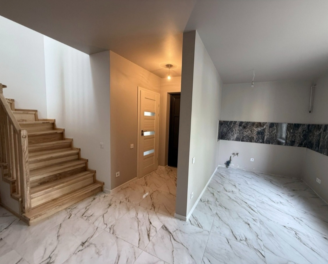 Jasne wnętrze domu z drewnianymi schodami i marmurową podłogą. Widoczne drzwi wewnętrzne z przeszkleniami i fragment ściany z dekoracyjnym pasem imitującym marmur.
