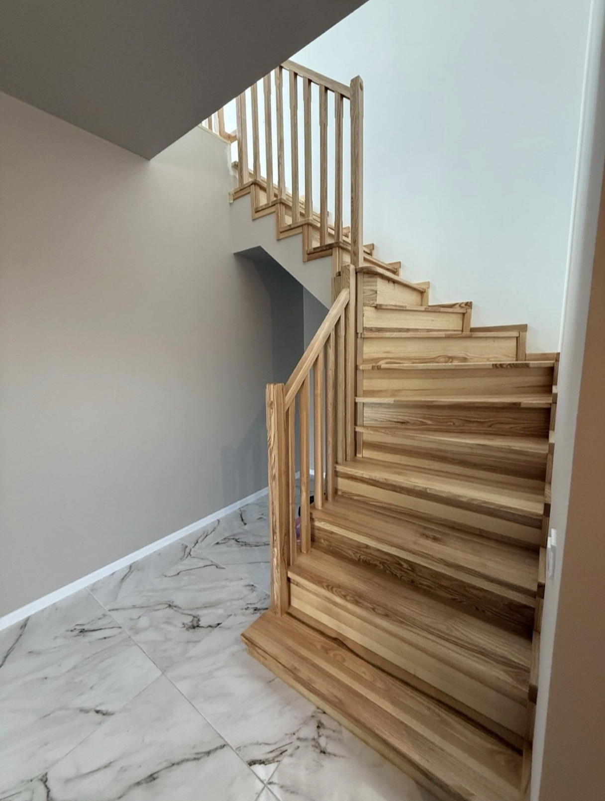 Jasne, drewniane schody z balustradą w nowoczesnym wnętrzu. Widoczne marmurowe płytki podłogowe i ściany w odcieniach szarości i bieli. Ujęcie z dołu, podkreślające konstrukcję.