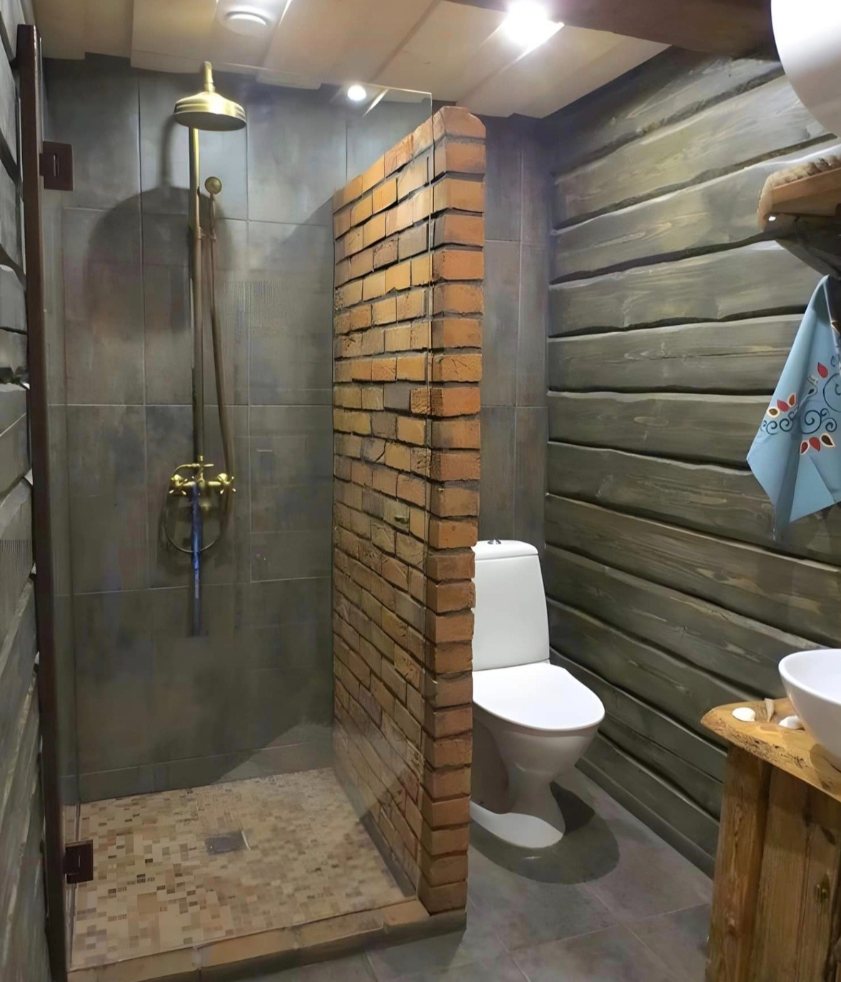 Unikalna łazienka z kabiną prysznicową oddzieloną ścianką z cegły, drewnianymi panelami ściennymi i złotą armaturą. Widoczna toaleta i umywalka na drewnianym blacie.
