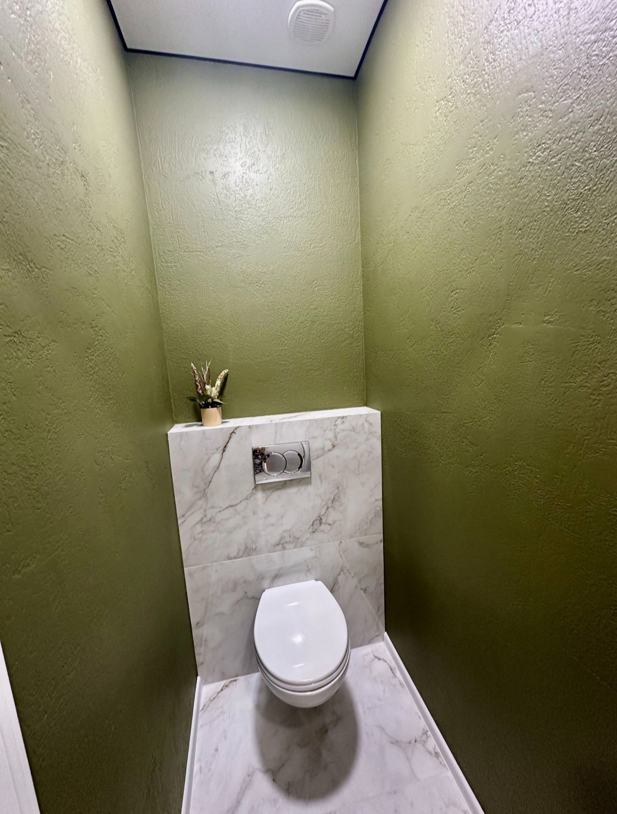 Wykończona toaleta w odcieniach zieleni i bieli. Ściany z fakturą, marmurowe płytki, wisząca miska WC i roślina w doniczce. Nowoczesny design, świeżo po remoncie.