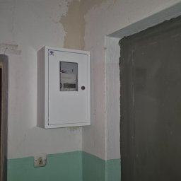 VOLT Usługi Elektryczne MAREK MICHURSKI - Biała skrzynka z licznikiem elektrycznym zamontowana na ścianie w korytarzu, widoczny fragment ściany pomalowanej na zielono i szare drzwi.