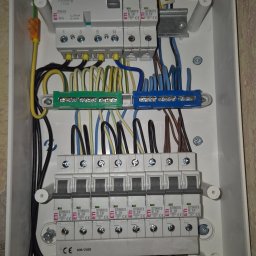 VOLT Usługi Elektryczne MAREK MICHURSKI - Rozdzielnica elektryczna w białej obudowie, widoczne bezpieczniki, przewody w kolorach: żółto-zielonym, niebieskim i czarnym. Instalacja po montażu, gotowa do użytku.