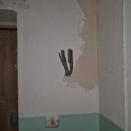 VOLT Usługi Elektryczne MAREK MICHURSKI - Wystające ze ściany przewody elektryczne, obok widoczny stary włącznik światła i ślady po naprawach tynku. Ujęcie z bliska, w tle fragment drzwi.