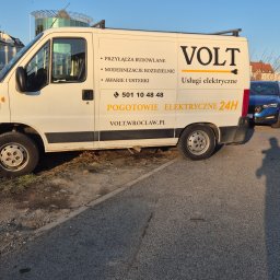 VOLT Usługi Elektryczne MAREK MICHURSKI - Biały samochód dostawczy z logo VOLT i informacjami o usługach elektrycznych, w tym pogotowiem elektrycznym 24h, zaparkowany na ulicy w słoneczny dzień.