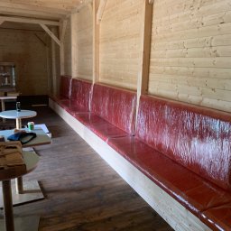 ZAKŁAD USŁUGOWO PRODUKCYJNY WOJCIECH SULKOWSKI - Długa, czerwona sofa z tworzywa sztucznego w drewnianej altanie. Widoczne stoliki kawowe. Sofa wykonana na wymiar, zabezpieczona folią ochronną.