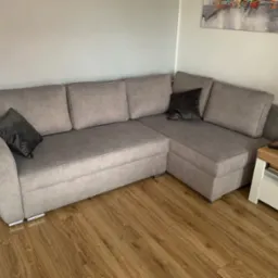 Szary narożnik z poduszkami w salonie, widok z boku. Narożnik stoi na drewnianej podłodze, obok niego stolik. Nad nim obraz na ścianie.