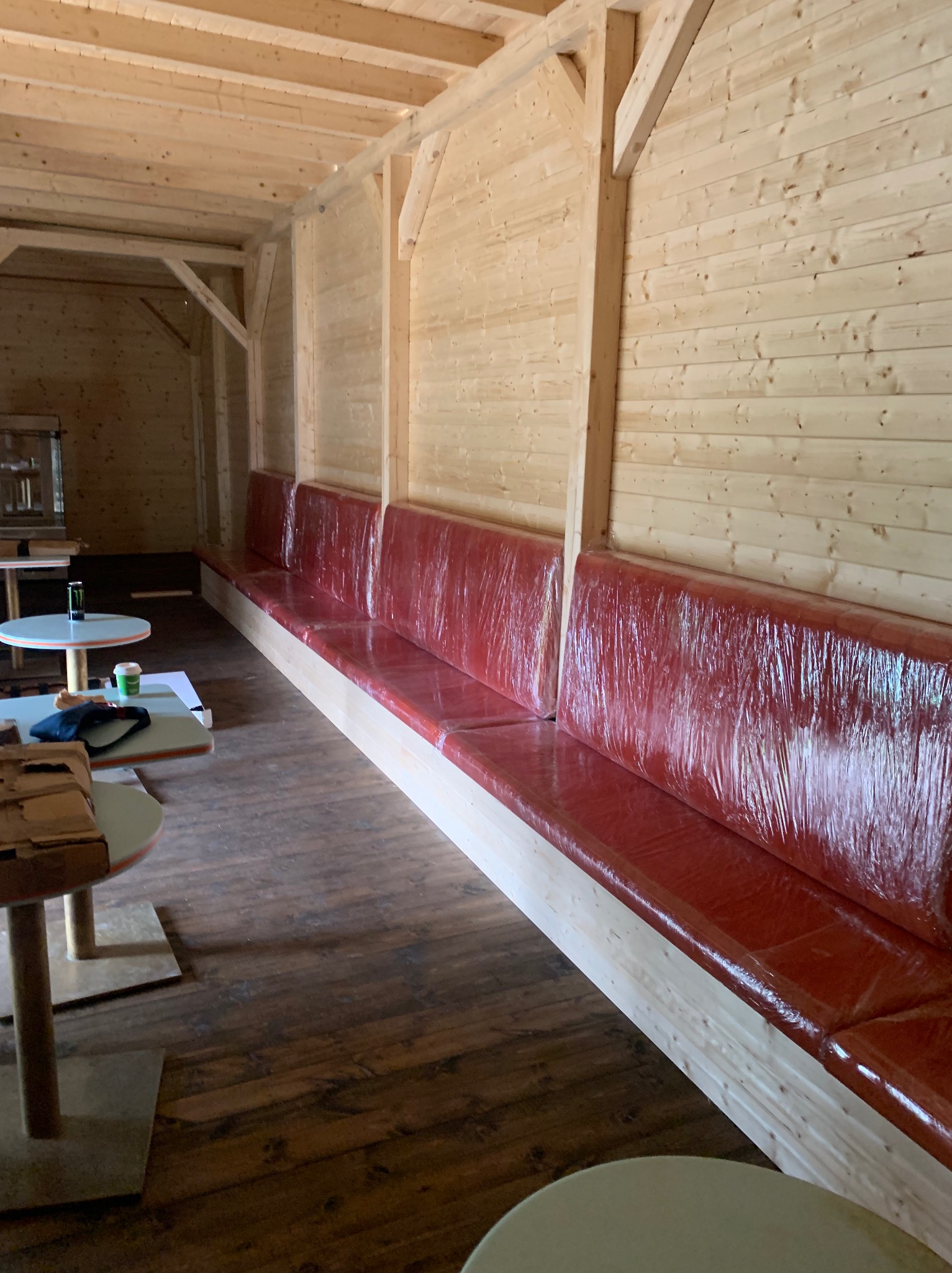 Długa, czerwona sofa z tworzywa sztucznego w drewnianej altanie. Widoczne stoliki kawowe. Sofa wykonana na wymiar, zabezpieczona folią ochronną.