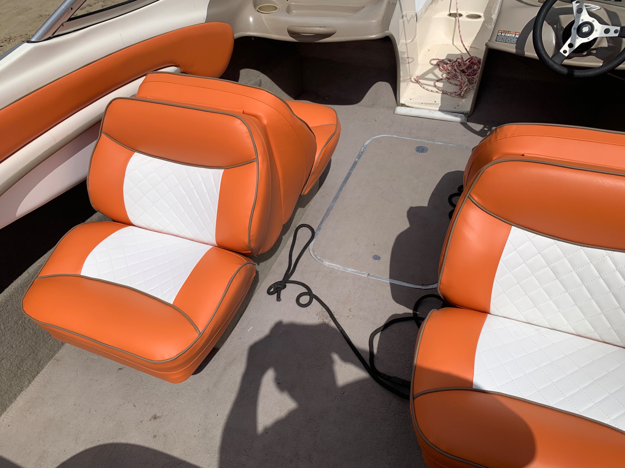 Tapicerowane, pomarańczowo-białe siedzenia w łodzi motorowej z czarną liną. Widoczny fragment kokpitu i kierownica. Czyste, jasne oświetlenie.