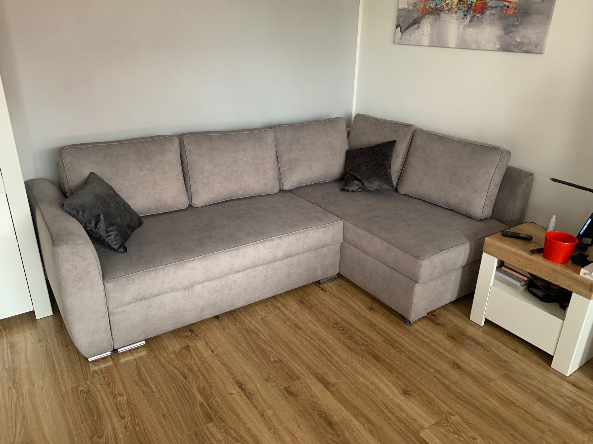 Szary narożnik z poduszkami w salonie, widok z boku. Narożnik stoi na drewnianej podłodze, obok niego stolik. Nad nim obraz na ścianie.