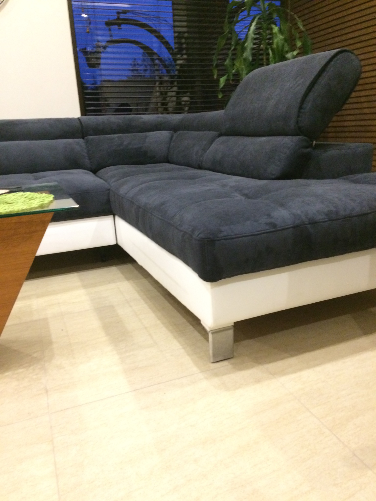 Szary narożnik z podnoszonym zagłówkiem, białą podstawą i metalowymi nóżkami stoi na podłodze z jasnych płytek. W tle okno z żaluzjami i roślina doniczkowa.