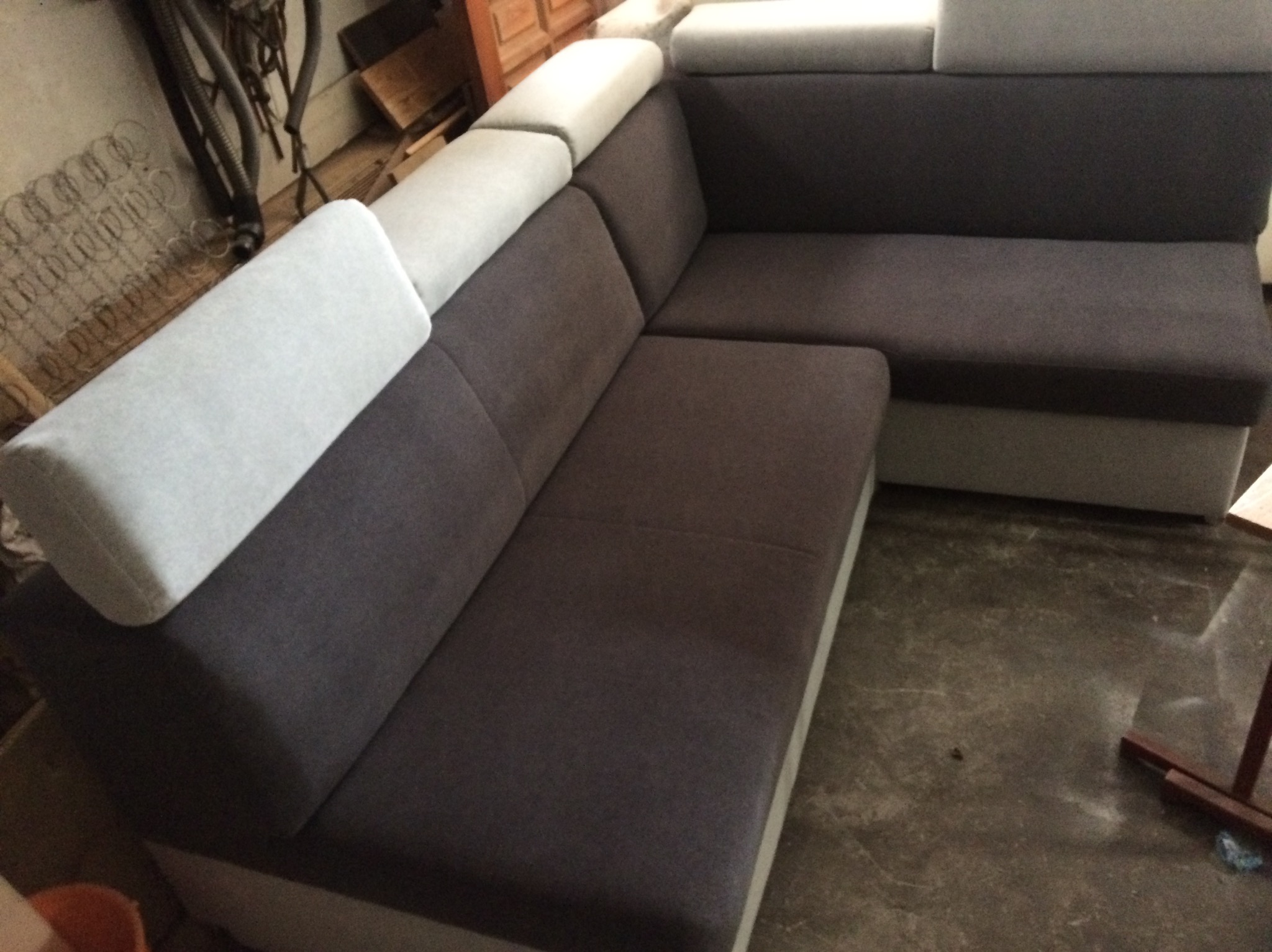 Szary narożnik z jasnymi zagłówkami, widoczny w warsztacie. Ujęcie z boku eksponuje detale tapicerki i kształt mebla. W tle narzędzia i materiały.