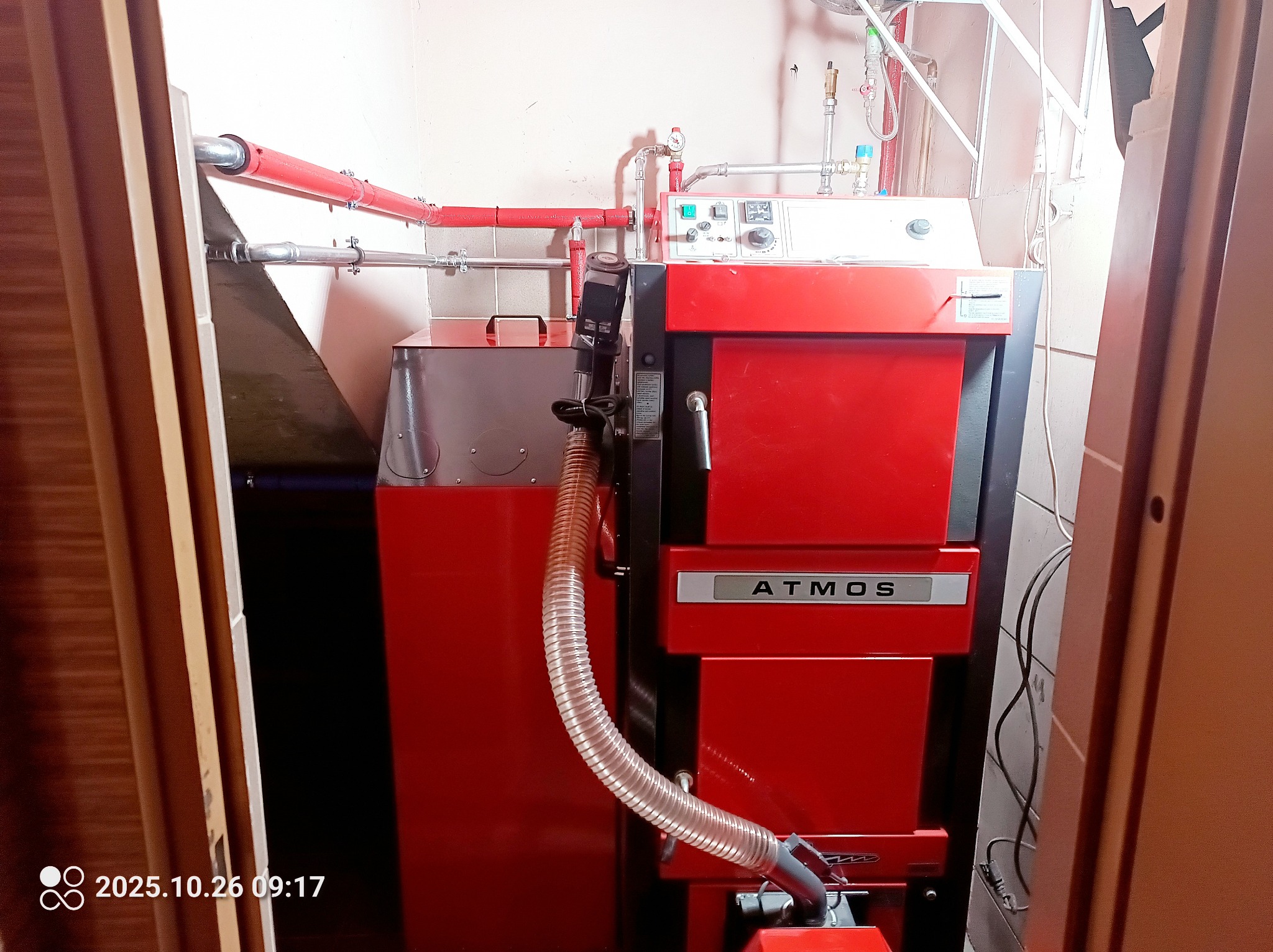 Czerwony piec CO marki Atmos z podłączonym przewodem, widoczny w kotłowni z rurami instalacji grzewczej. Ujęcie z bliska, podkreślające detale instalacji.