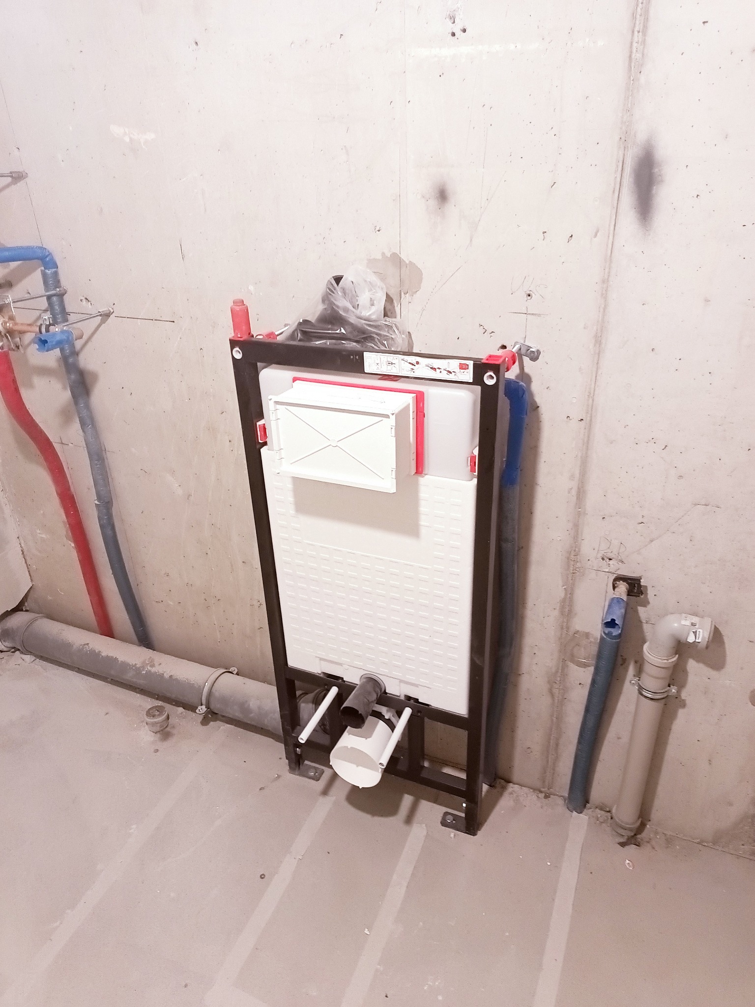 Stelaż podtynkowy WC z widocznymi przyłączami wodno-kanalizacyjnymi na surowej ścianie. Widoczne rury w kolorze niebieskim i czerwonym. Szara podłoga.