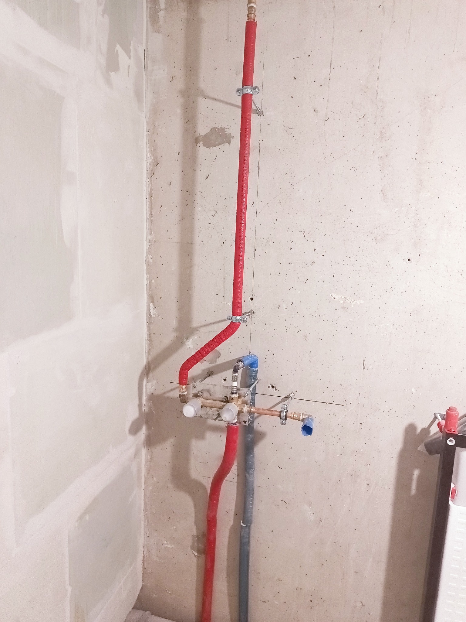 Fragment instalacji hydraulicznej z czerwonymi rurami w izolacji, zamontowanej na surowej ścianie w trakcie prac wykończeniowych. Widoczne elementy miedziane i niebieskie.