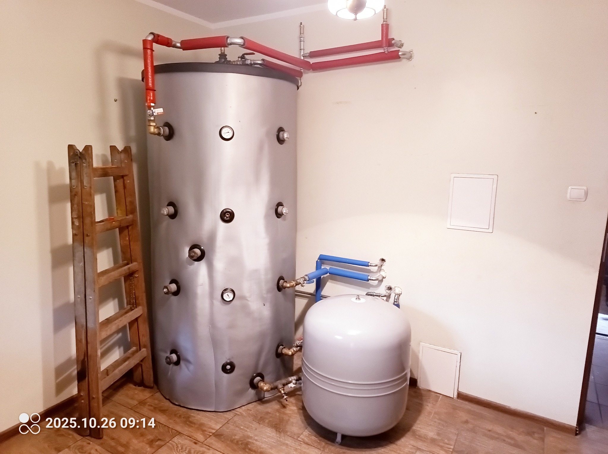 Szary, pionowy zbiornik akumulacyjny z czerwonymi i niebieskimi rurami, obok drabina. Element instalacji grzewczej w pomieszczeniu.