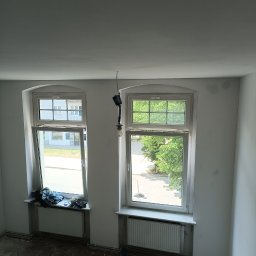 Ivan Husiev - Pokój w trakcie prac przygotowawczych do malowania: ściany świeżo zagruntowane, podłoga zabezpieczona przed zabrudzeniem, wisząca żarówka na kablu, widok z góry na okna z grzejnikami.