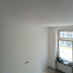 Ivan Husiev - Pokój w trakcie malowania na biało, widoczne ślady farby na podłodze, odsłonięte gniazdka elektryczne i okno z widokiem na budynek mieszkalny.