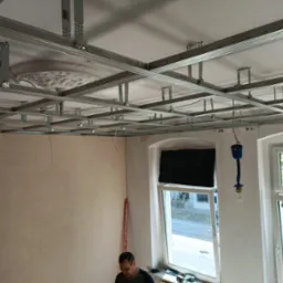 Montaż metalowej konstrukcji sufitu podwieszanego z widoczną rozetą dekoracyjną, ściany przygotowane do dalszych prac wykończeniowych, widok przez okno na ulicę.