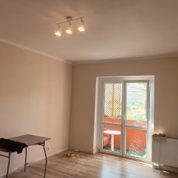 Ivan Husiev - Pomalowane na beżowo puste pomieszczenie z balkonem, widoczny stolik, grzejnik z butelką płynu do czyszczenia i lampa sufitowa z trzema kloszami.