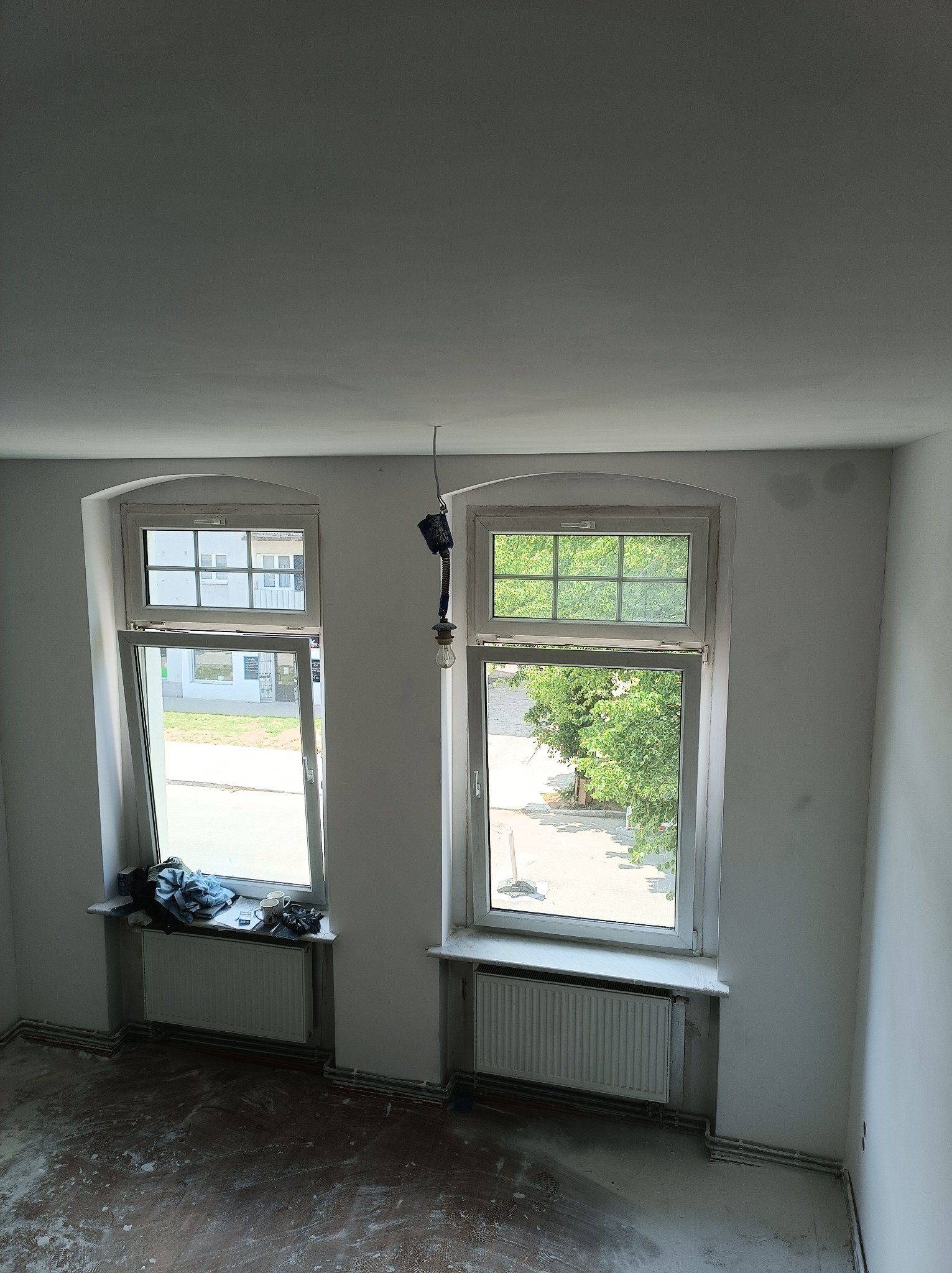 Pokój w trakcie prac przygotowawczych do malowania: ściany świeżo zagruntowane, podłoga zabezpieczona przed zabrudzeniem, wisząca żarówka na kablu, widok z góry na okna z grzejnikami.