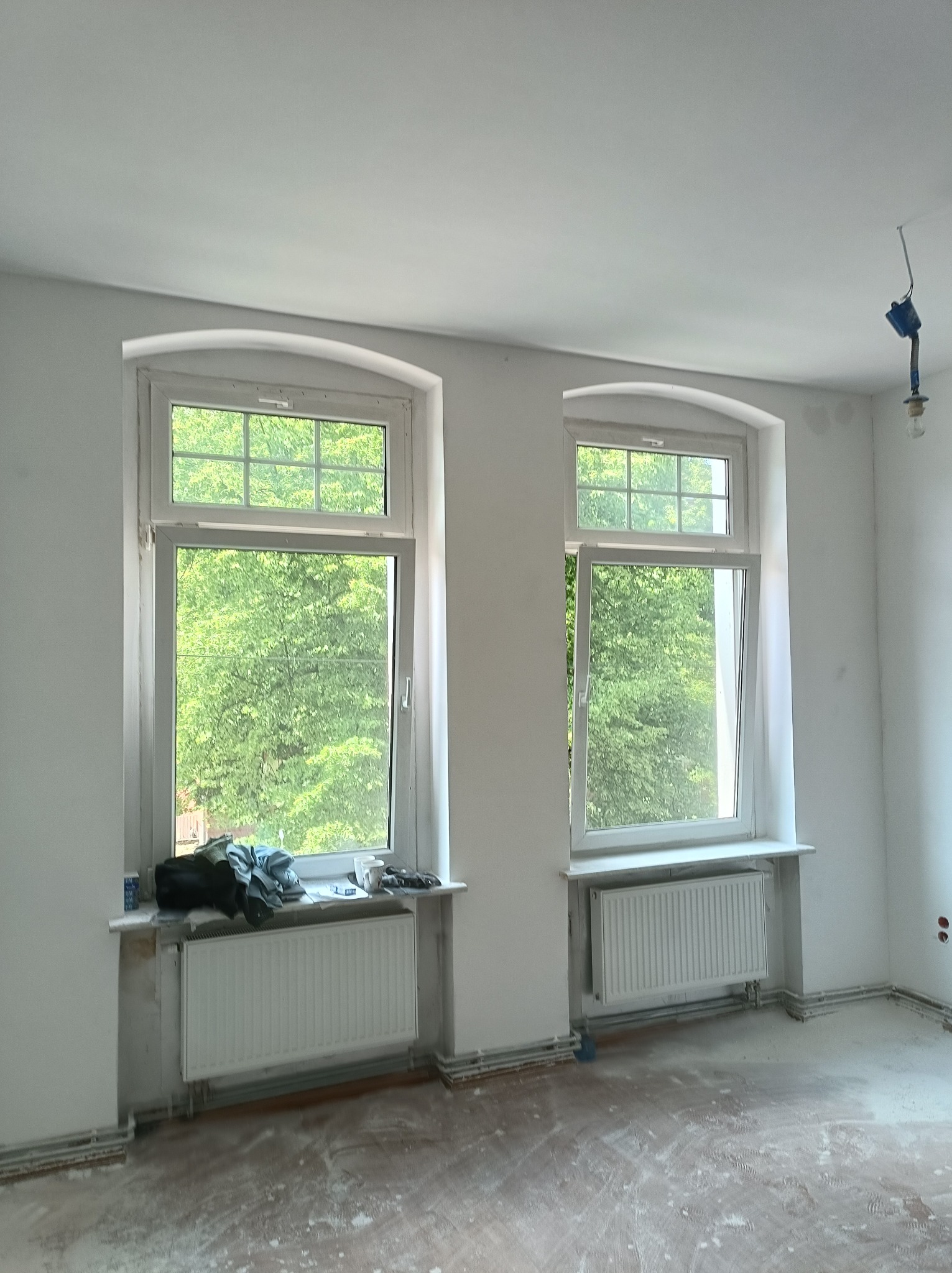 Pokój po wstępnych pracach przygotowawczych, widoczne dwa okna z białymi ramami, grzejniki, ślady po pracach na podłodze, gołe ściany z widocznymi punktami elektrycznymi.