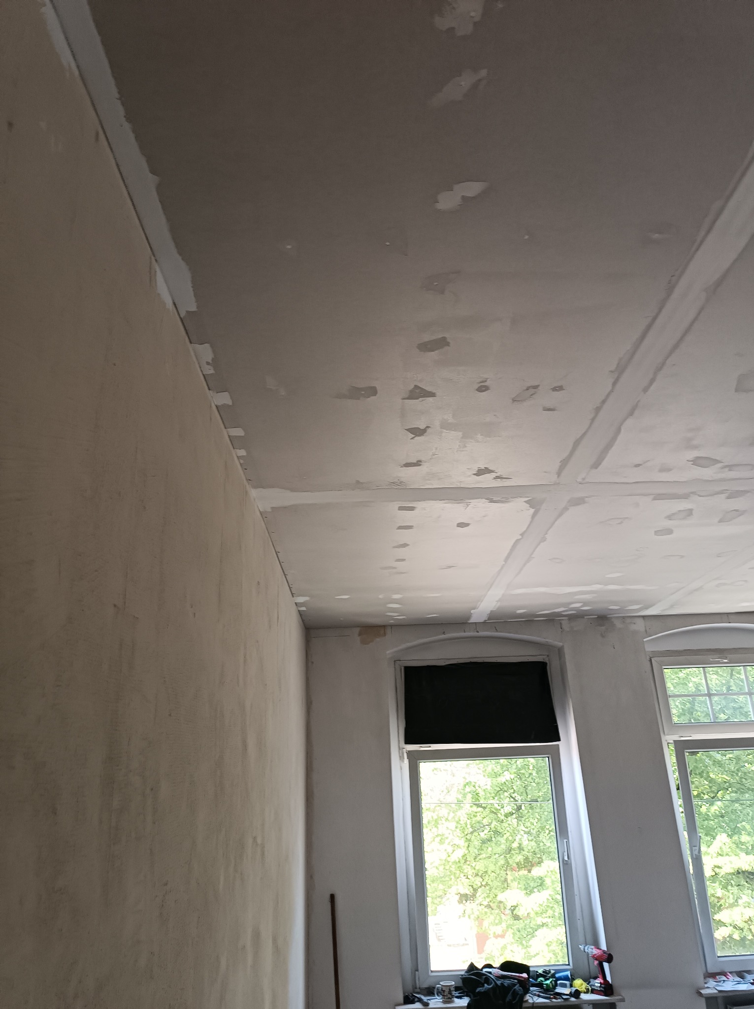 Przygotowana do gładzi ściana z widocznymi łączeniami płyt karton-gips na suficie, narzędzia i akcesoria na parapecie okiennym.