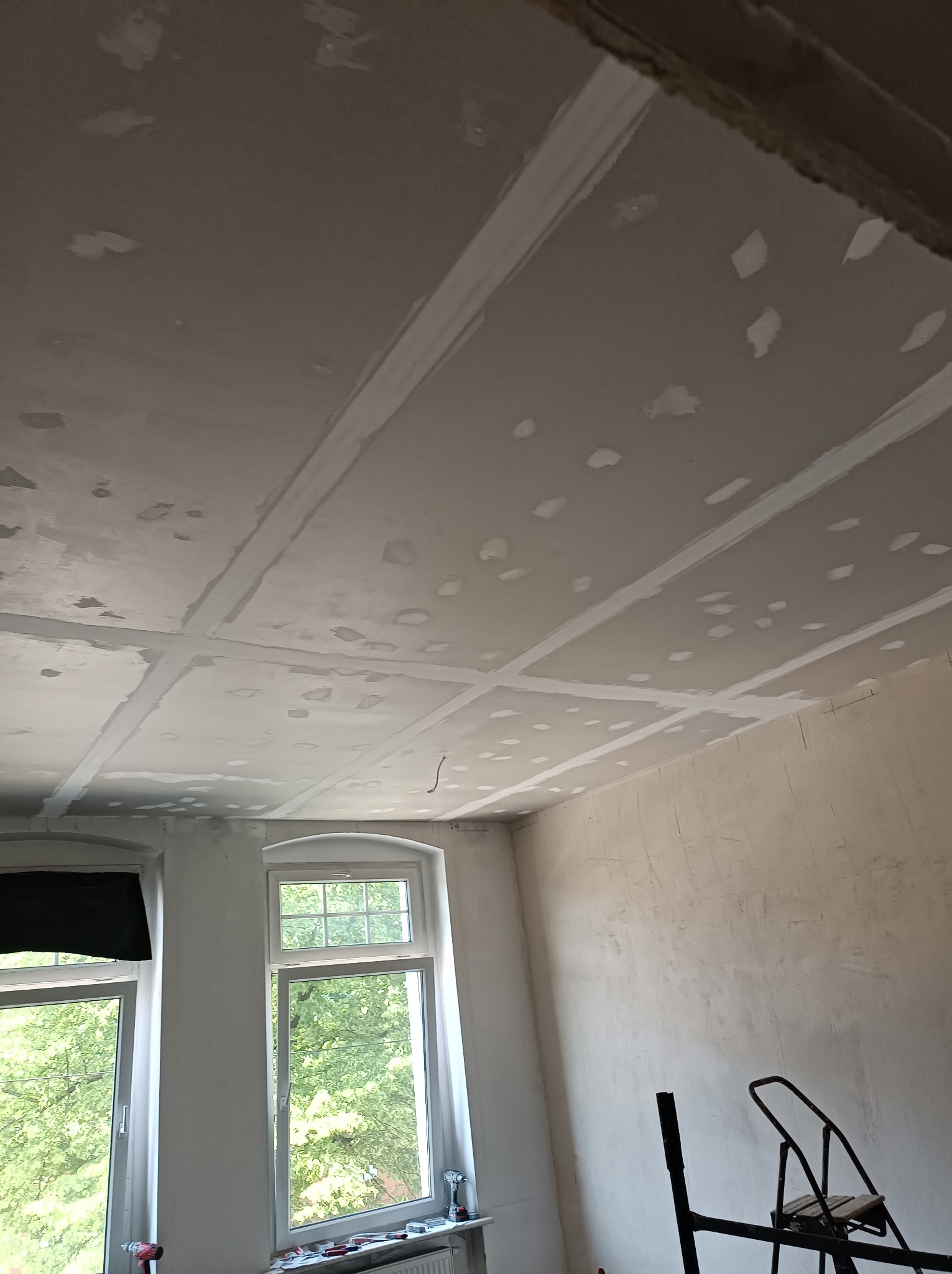 Sufit z płyt kartonowo-gipsowych, szpachlowany na łączeniach, widoczne ślady po wkrętach, ściana w trakcie szpachlowania, fragment drabiny w rogu pokoju, dwa okna z widokiem na zieleń.