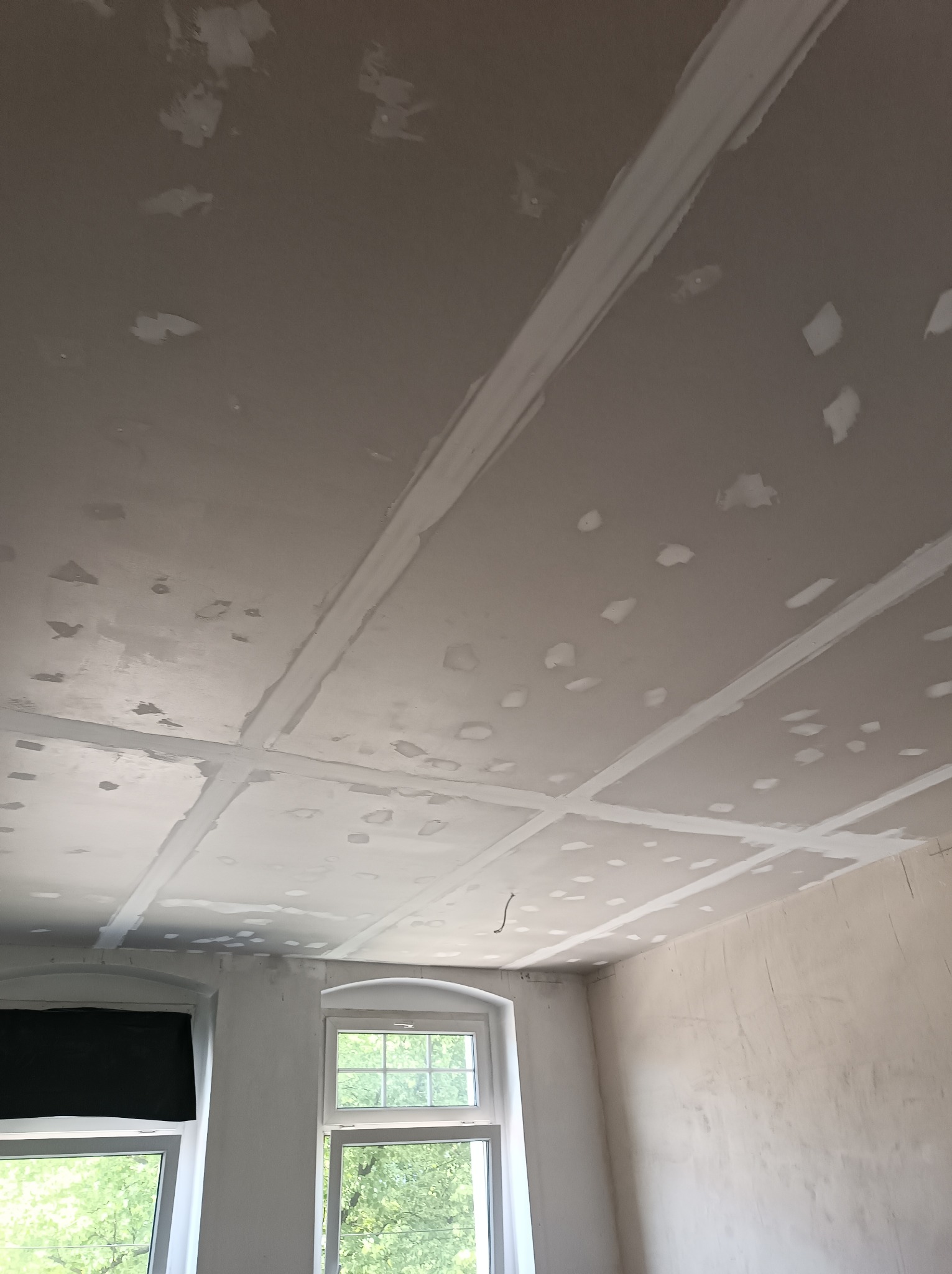 Przygotowanie sufitu pod gładzie: widoczne łączenia płyt kartonowo-gipsowych zaszpachlowane, ślady po wkrętach również zaszpachlowane, sufit w trakcie prac, dwa okna w tle z widokiem na zieleń.