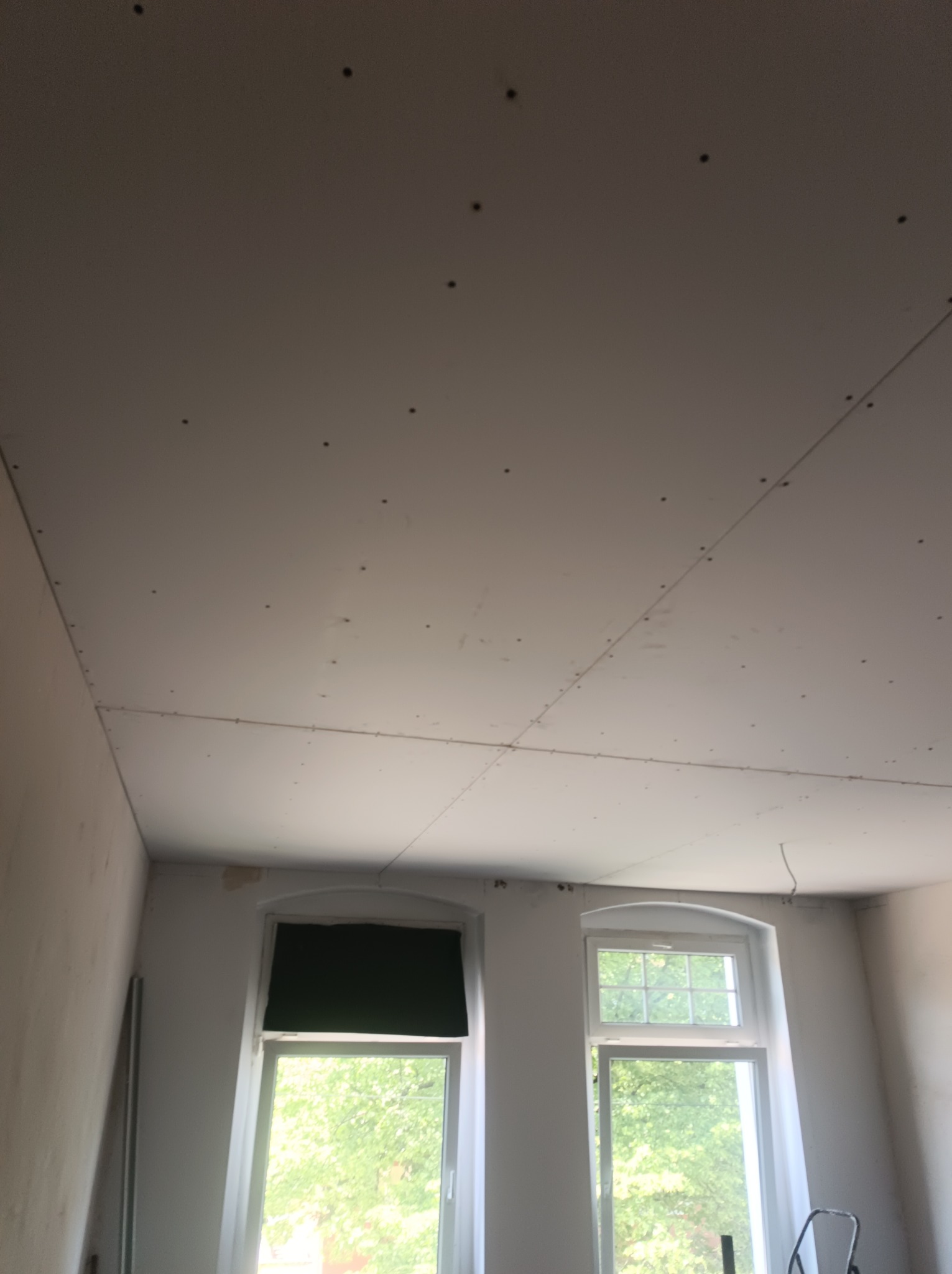 Sufit z płyt kartonowo-gipsowych, widoczne łączenia i ślady po wkrętach, dwa okna z widokiem na zieleń na ścianie w tle.