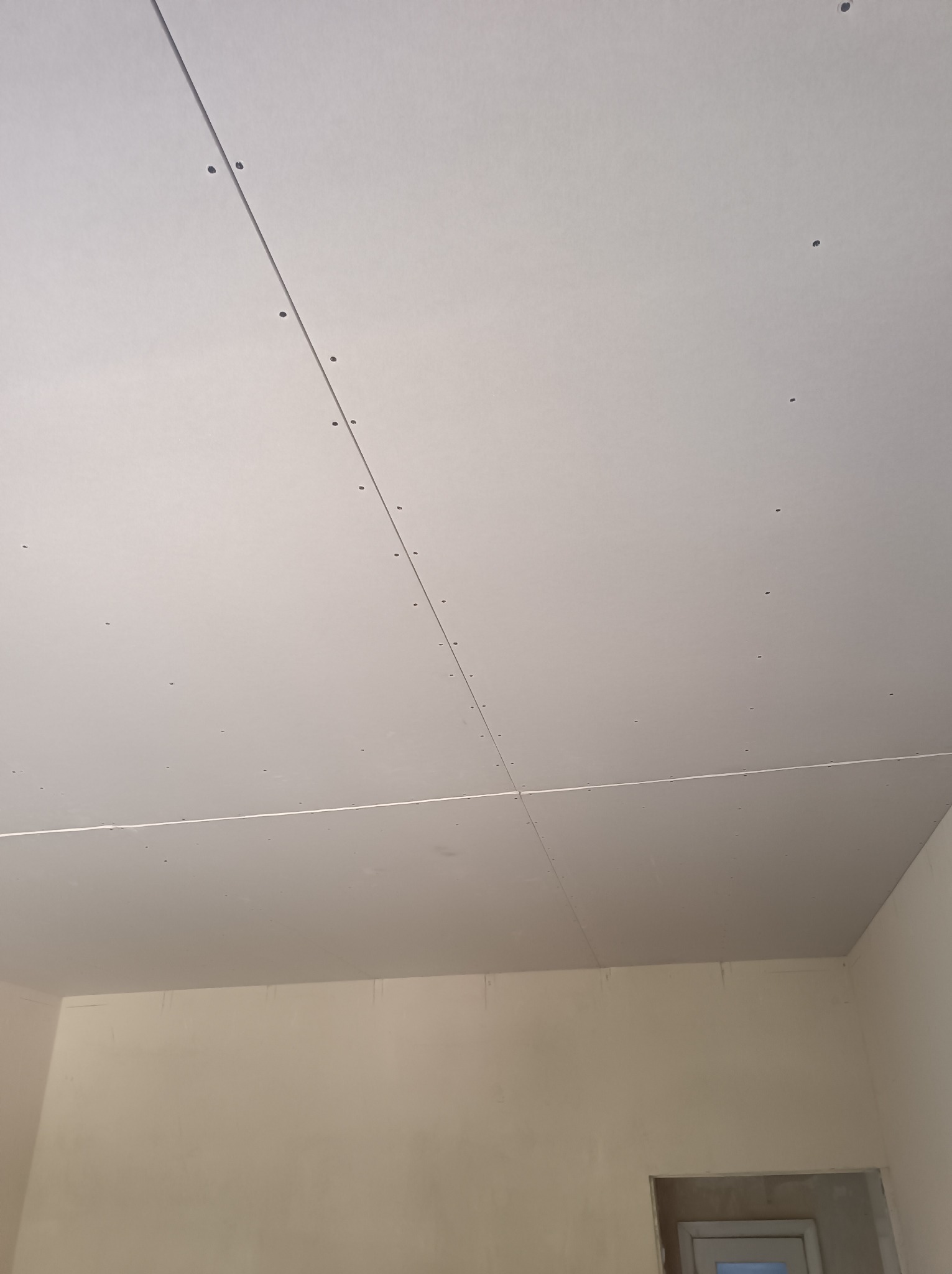 Zamocowany sufit z płyt kartonowo-gipsowych, widoczne łączenia i ślady po wkrętach, ściany przygotowane do malowania, widoczny fragment ościeżnicy drzwiowej.