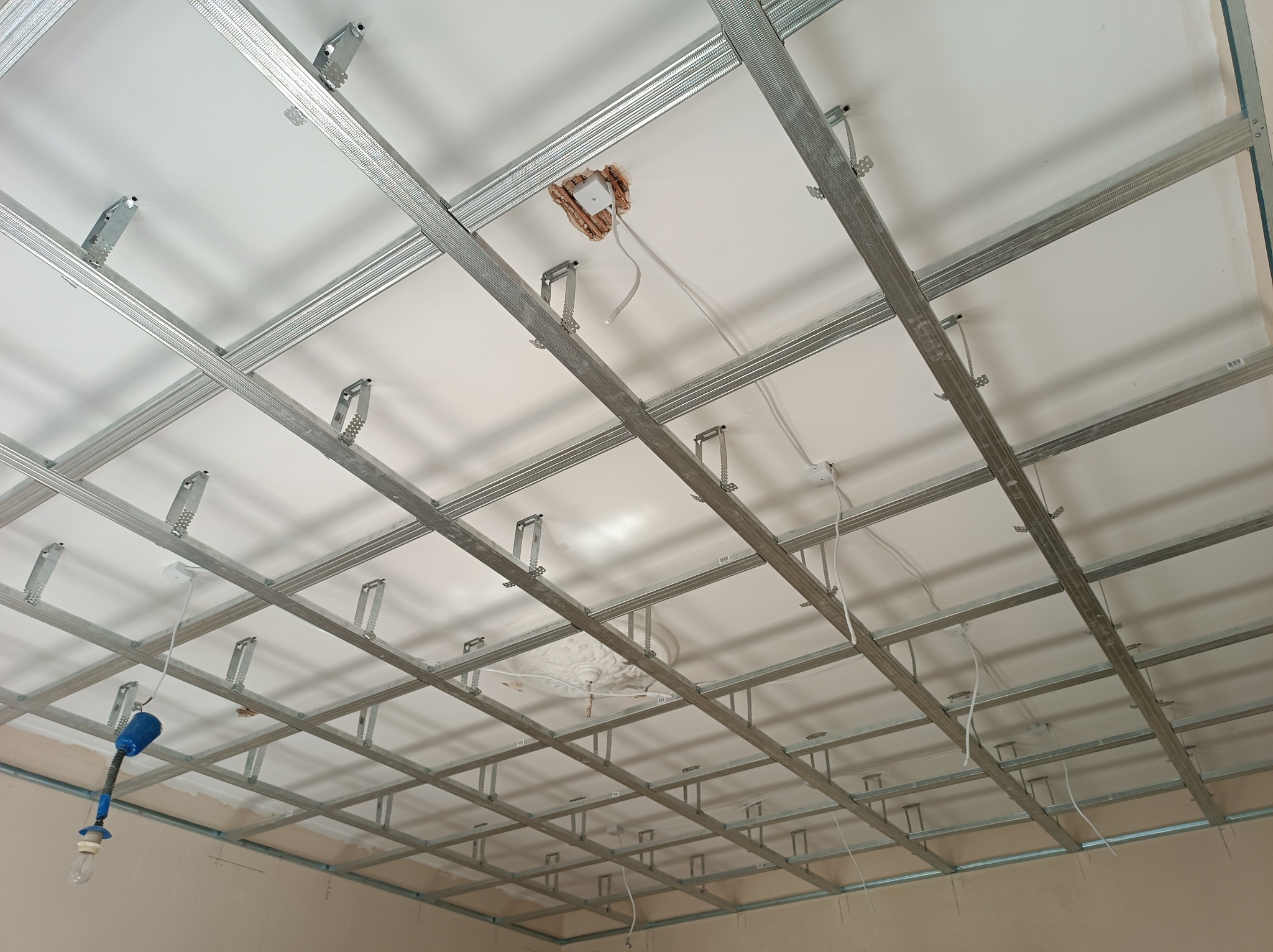 Sufit podwieszany z metalową konstrukcją w trakcie montażu, widoczne przewody elektryczne i otwór na oświetlenie.