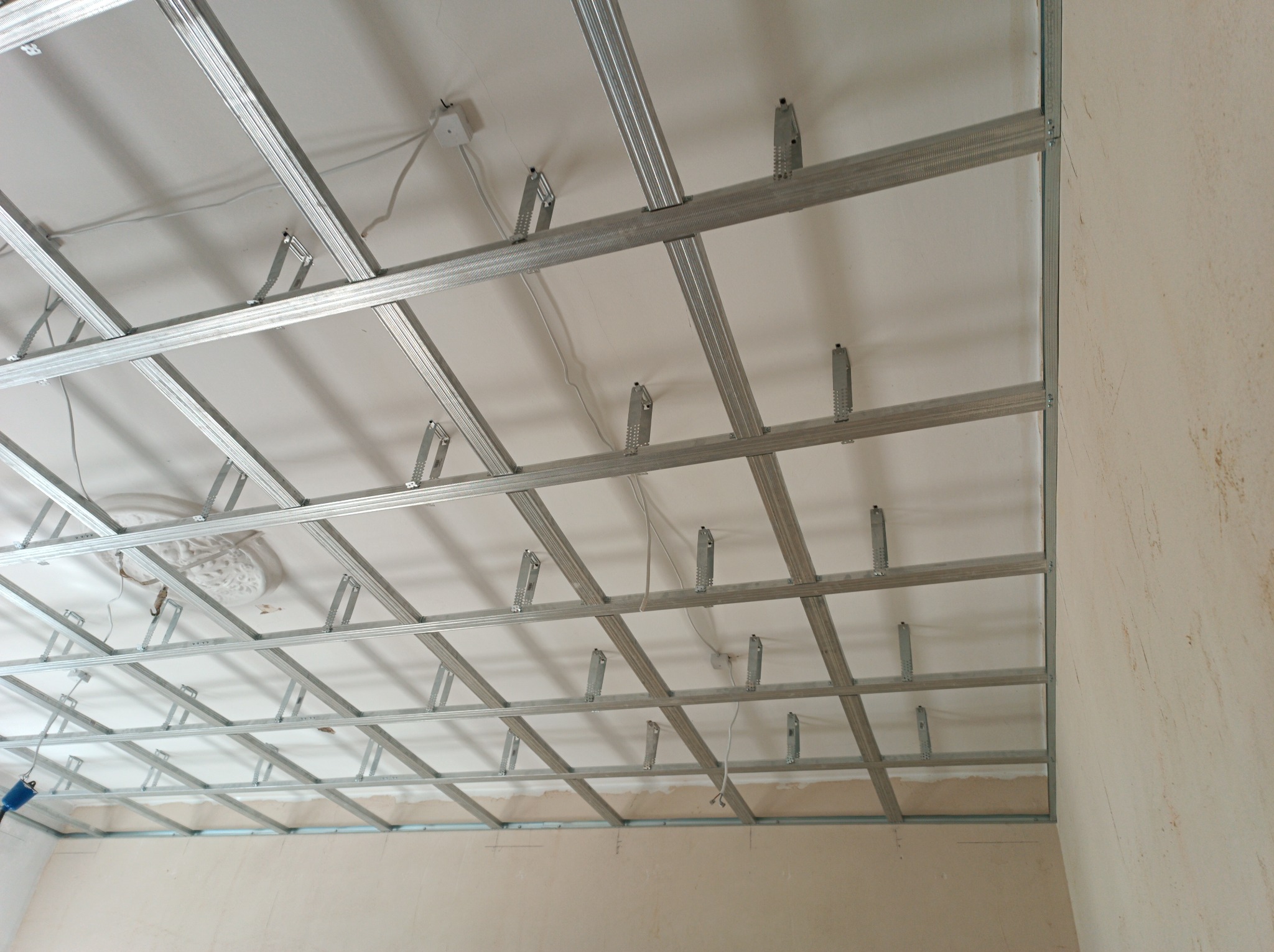 Metalowa konstrukcja sufitu podwieszanego w trakcie montażu, widoczne profile i elementy mocujące, surowe ściany w tle.