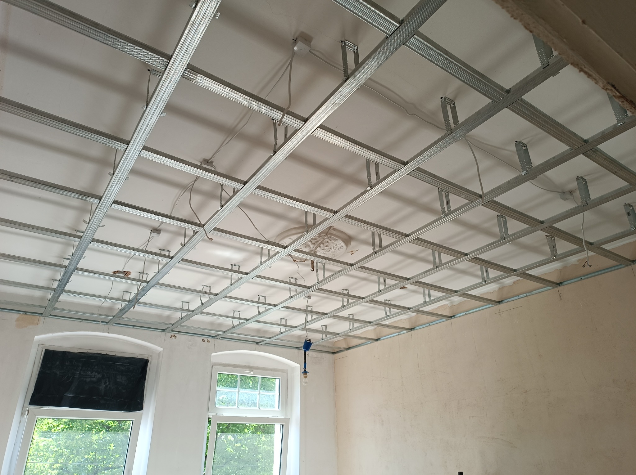 Stelaż sufitu podwieszanego z płyt karton-gips, widoczne profile stalowe i zawiesia, instalacja elektryczna w przestrzeni sufitu, okna zabezpieczone folią malarską.