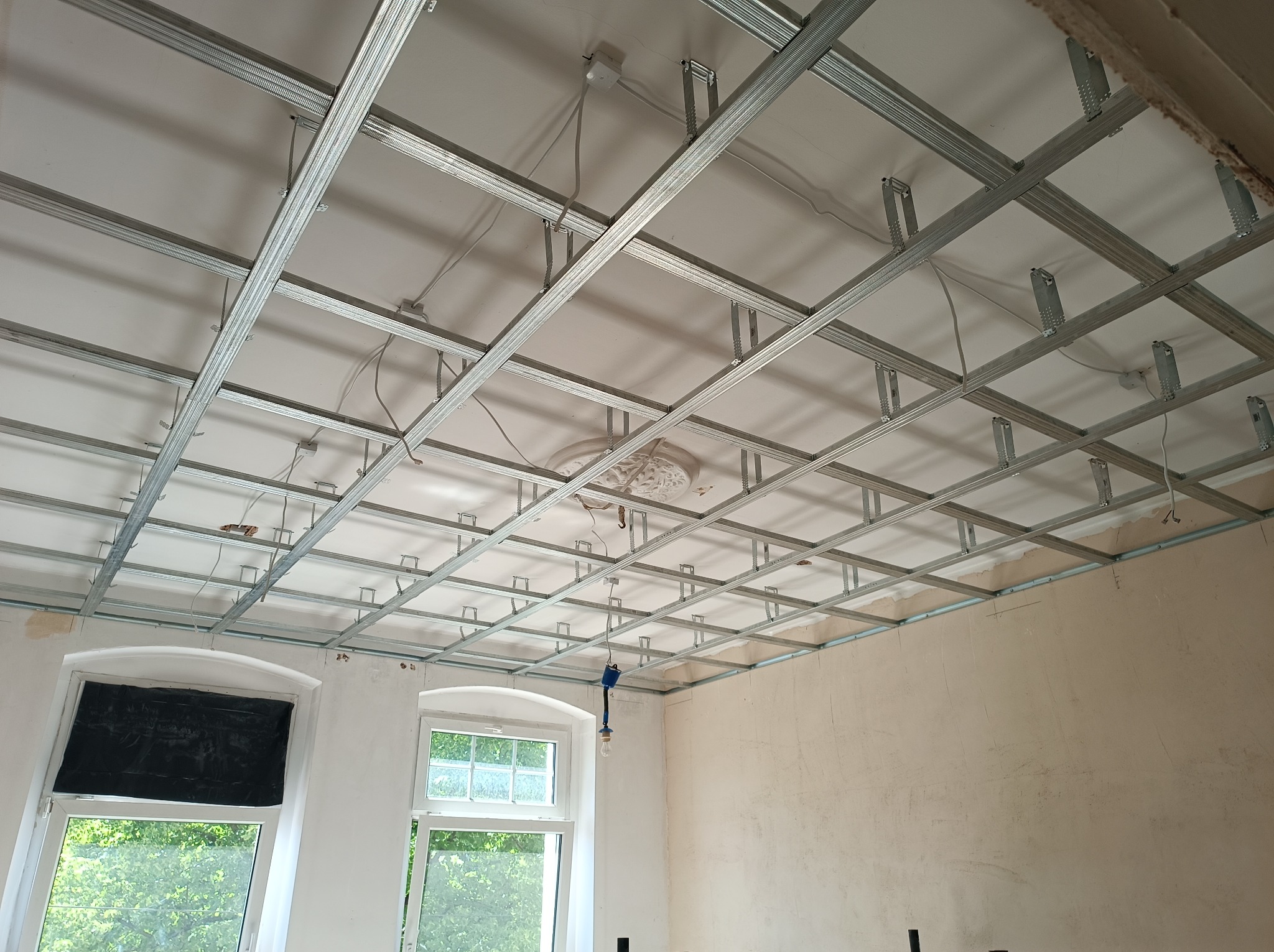 Stelaż sufitu podwieszanego z profili metalowych w trakcie montażu, widoczne okna z zasłoniętymi szybami i przewody elektryczne