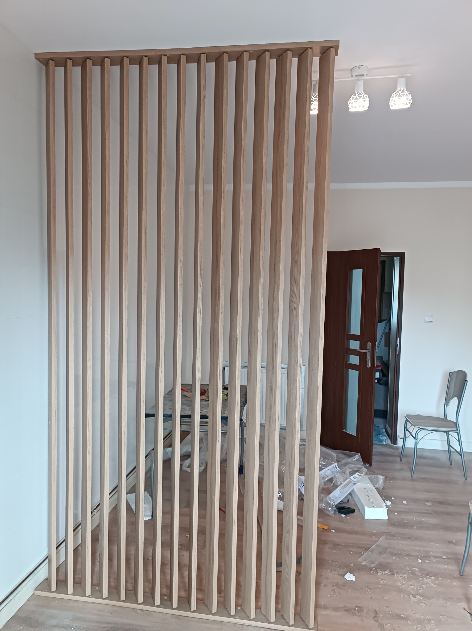 Drewniana ażurowa ścianka działowa z pionowych listew oddzielająca przestrzeń w mieszkaniu, widoczne elementy montażowe i resztki materiałów po instalacji.