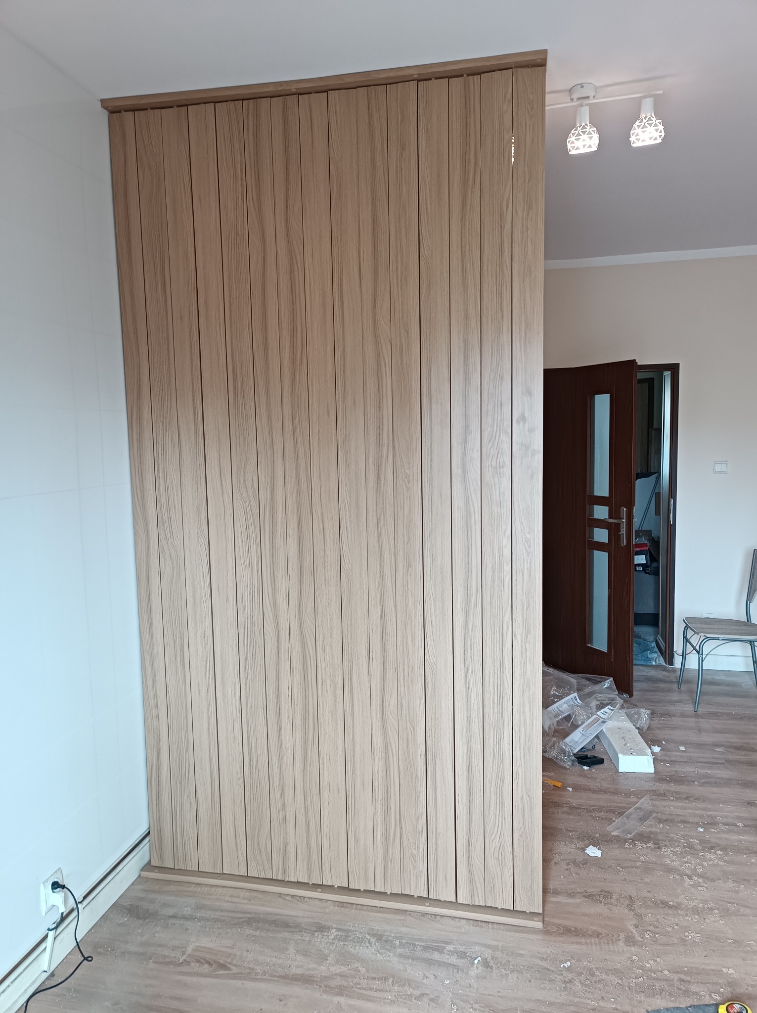 Montaż zabudowy meblowej z paneli drewnopodobnych, widoczne elementy mocujące i resztki materiałów na podłodze.