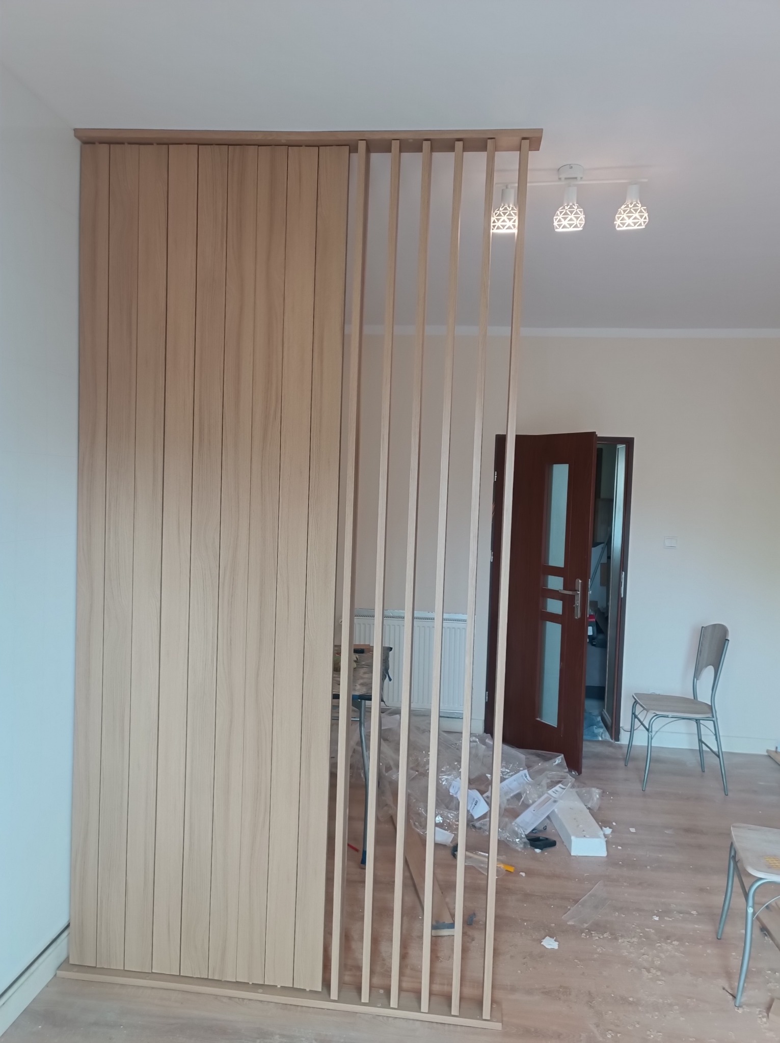 Drewniana ścianka działowa z pionowych listew, częściowo zabudowana, w jasnym wnętrzu w trakcie montażu, widoczne narzędzia i materiały.